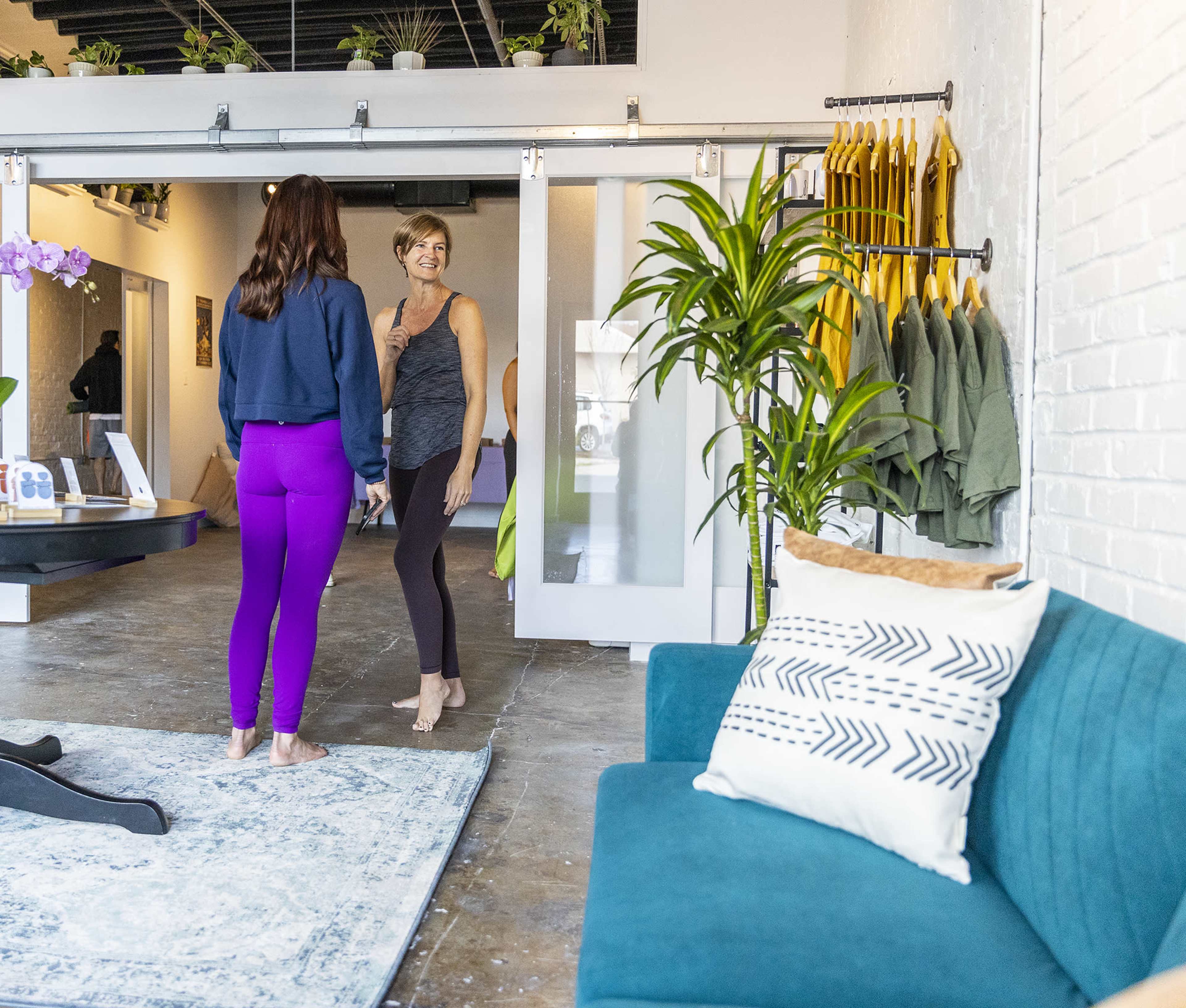 Two women are engaged in conversation in a spacious, modern room with plants and yoga attire displayed.