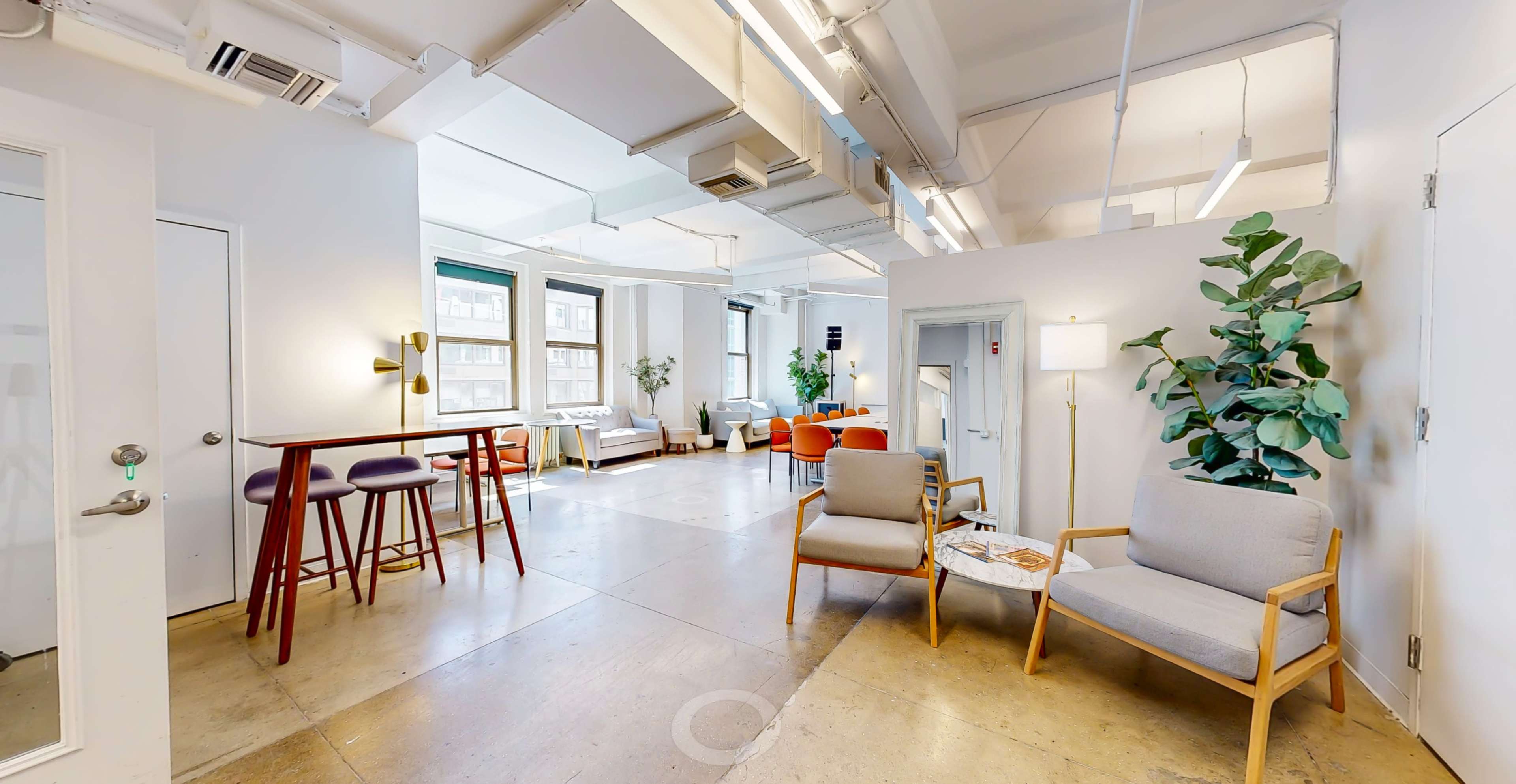 A modern open office space featuring seating areas with gray chairs, a small bar table, green plants, and large windows allowing natural light.