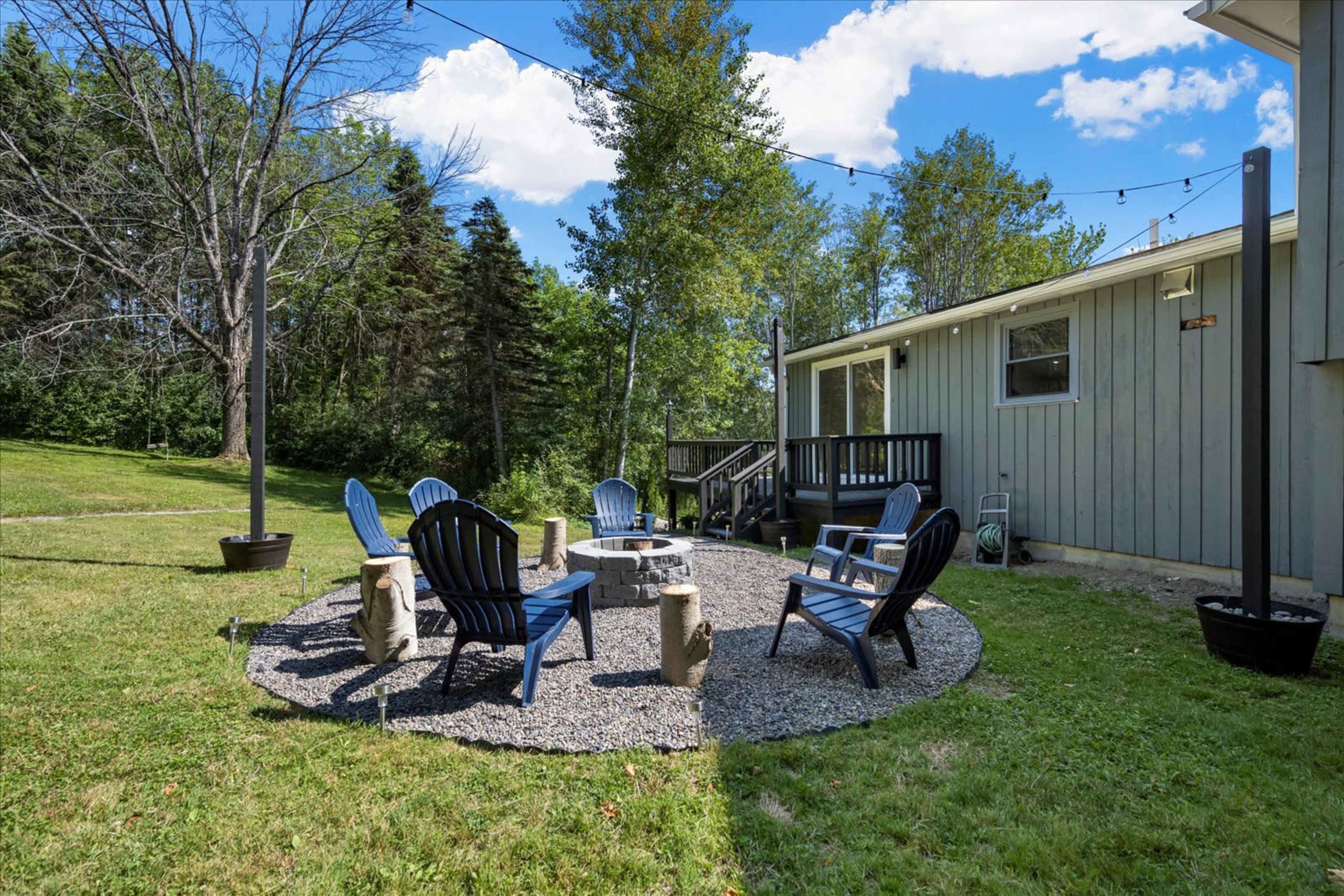 A circular seating area with blue chairs surrounds a stone fire pit in a grassy outdoor space next to a house.