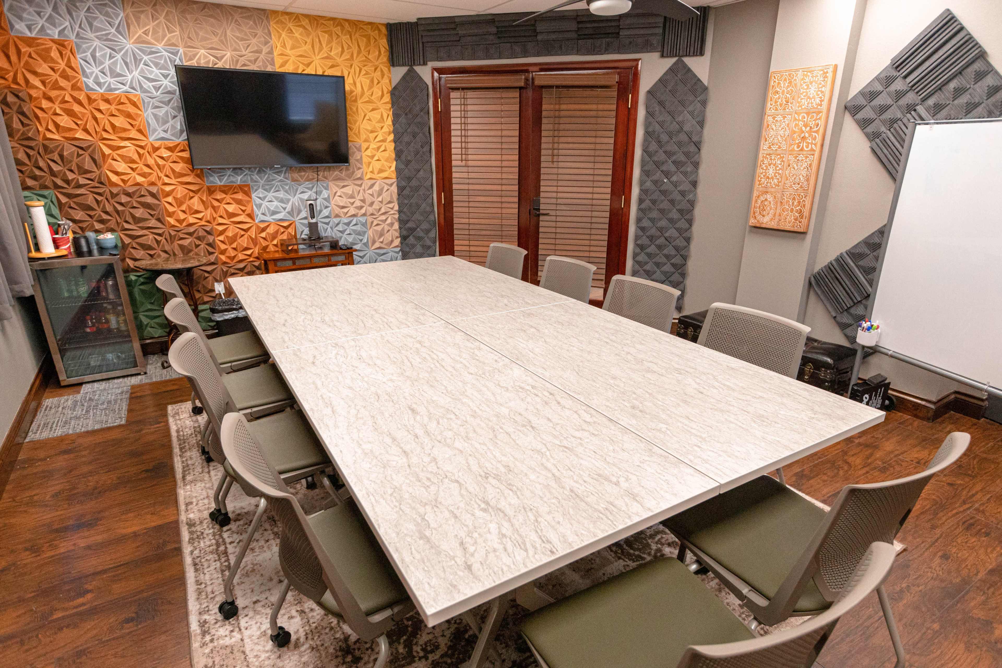 The image shows a modern conference room with a large table surrounded by chairs, and walls featuring textured acoustic panels and a television mounted on one side.