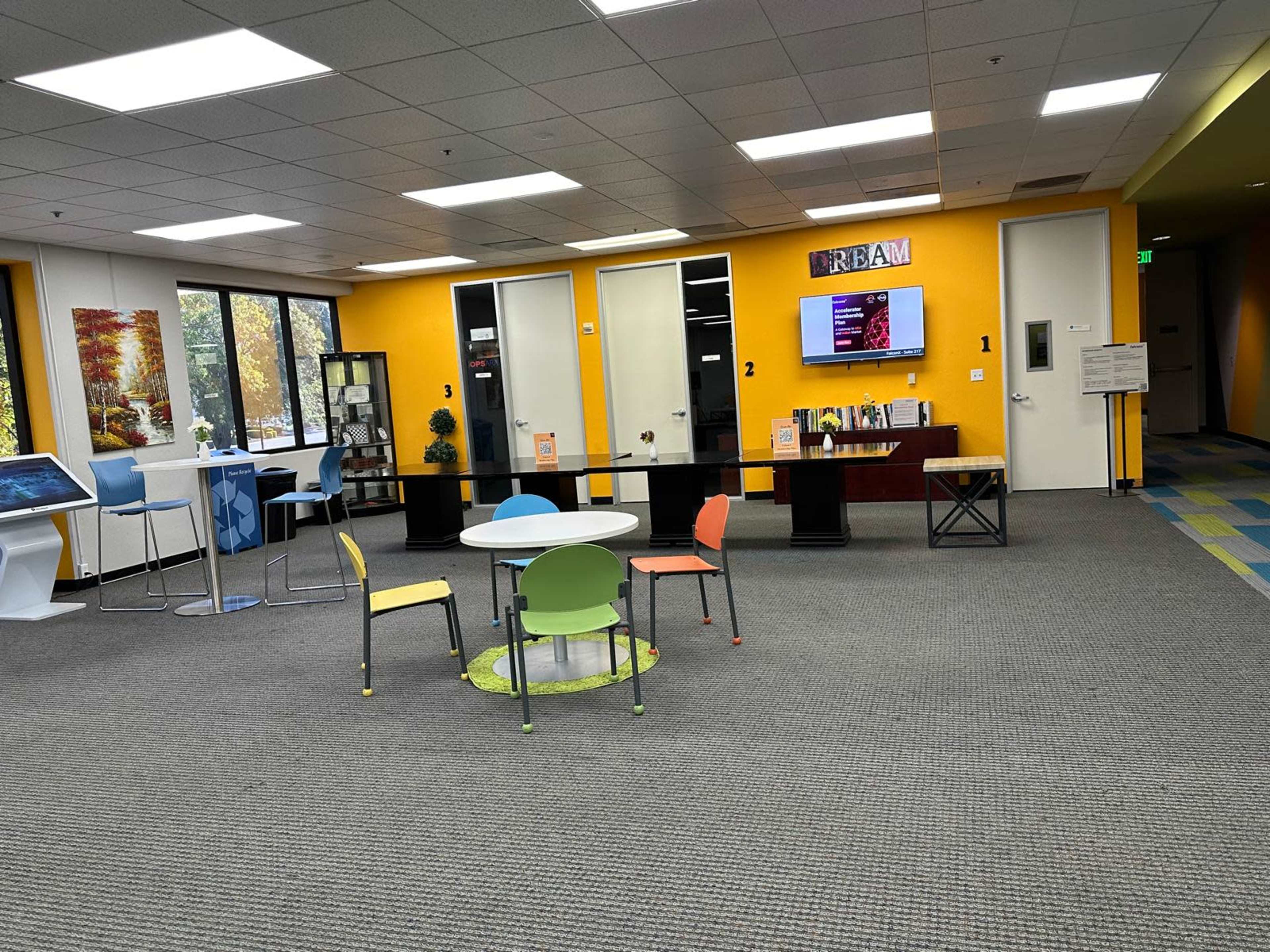 The image shows a brightly colored office space with a round table and colorful chairs, separated by partitions, featuring a TV display on the wall.