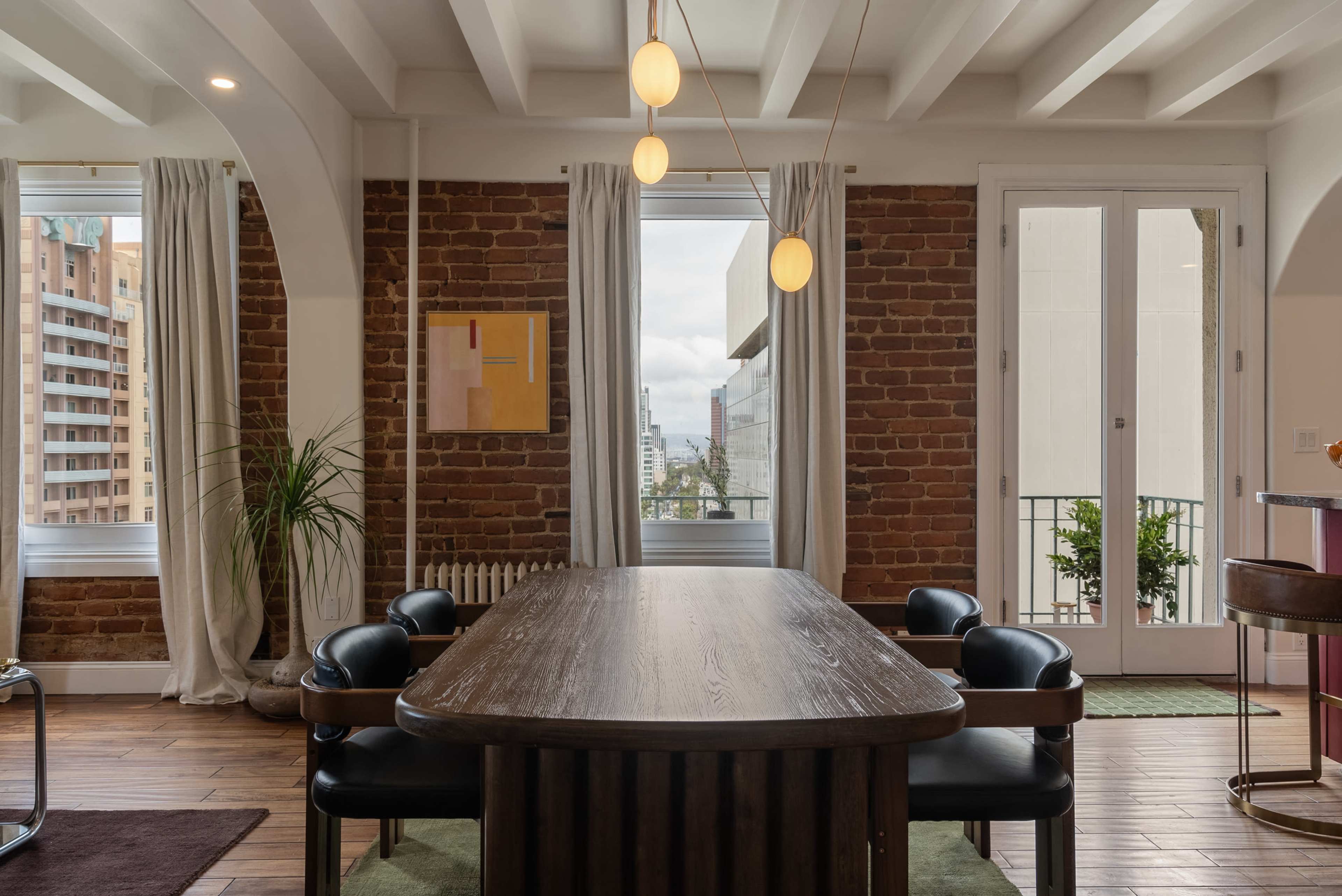 A dining area features a wooden table surrounded by black chairs, with large windows showcasing a view of a cityscape.