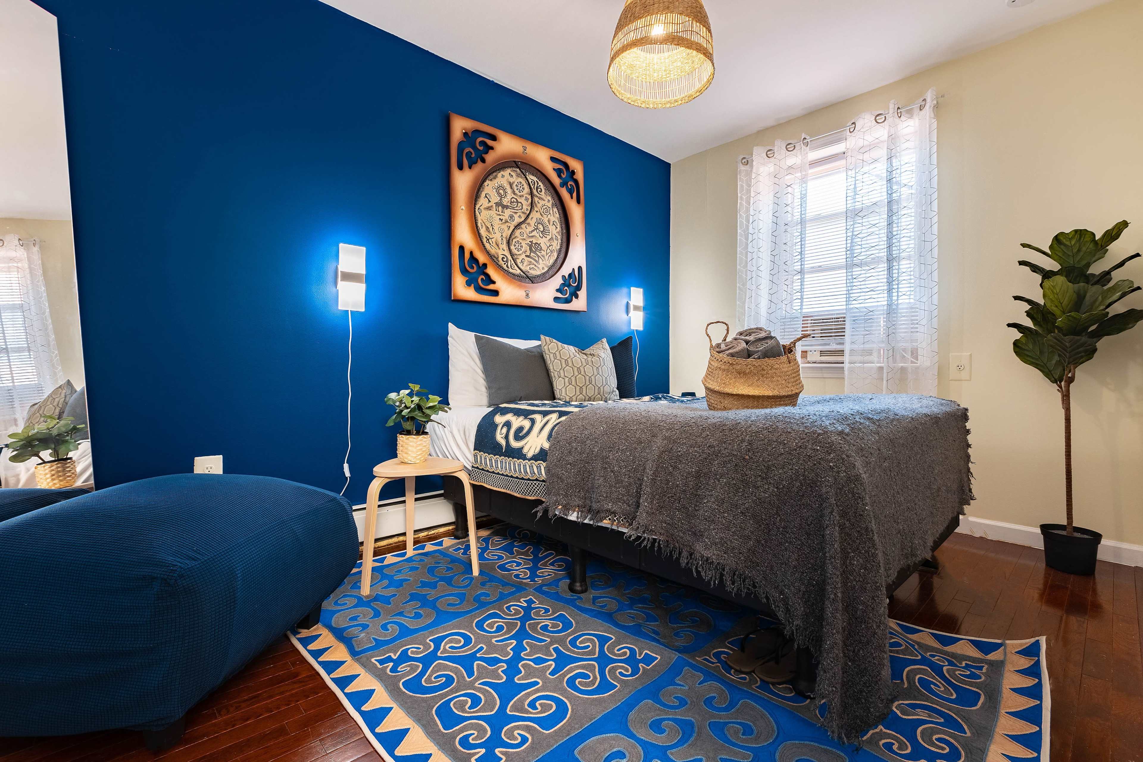 A cozy bedroom features a blue accent wall, a bed with a gray blanket, a decorative round wall piece, a plant, and a blue area rug.