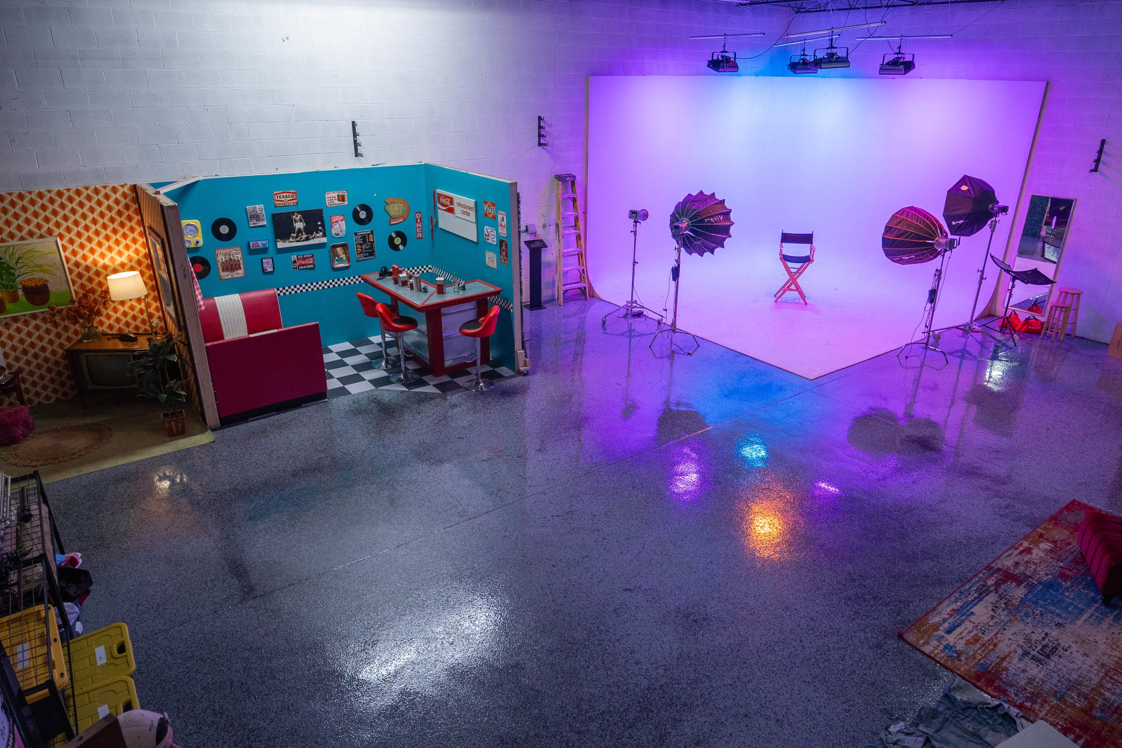 The image shows a spacious studio with a colorful adorned wall, a dining set, and a brightly lit white backdrop featuring studio lights.