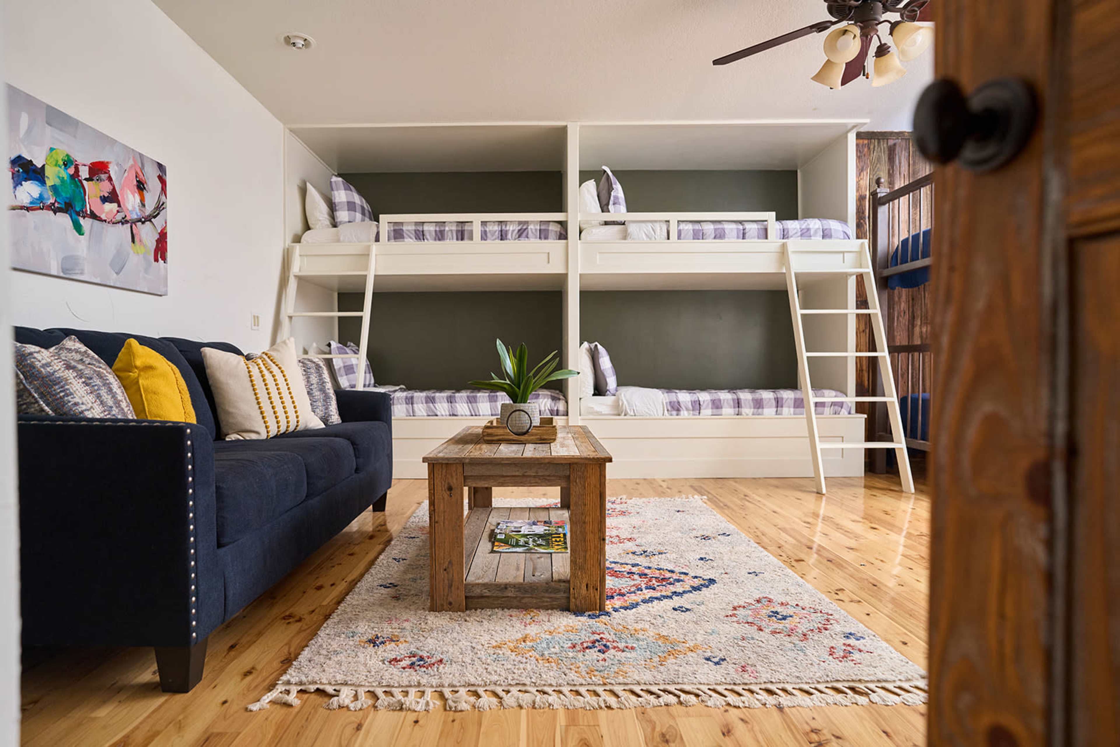 The image shows a cozy room featuring a blue sofa, a wooden coffee table, and two sets of bunk beds against a wall.