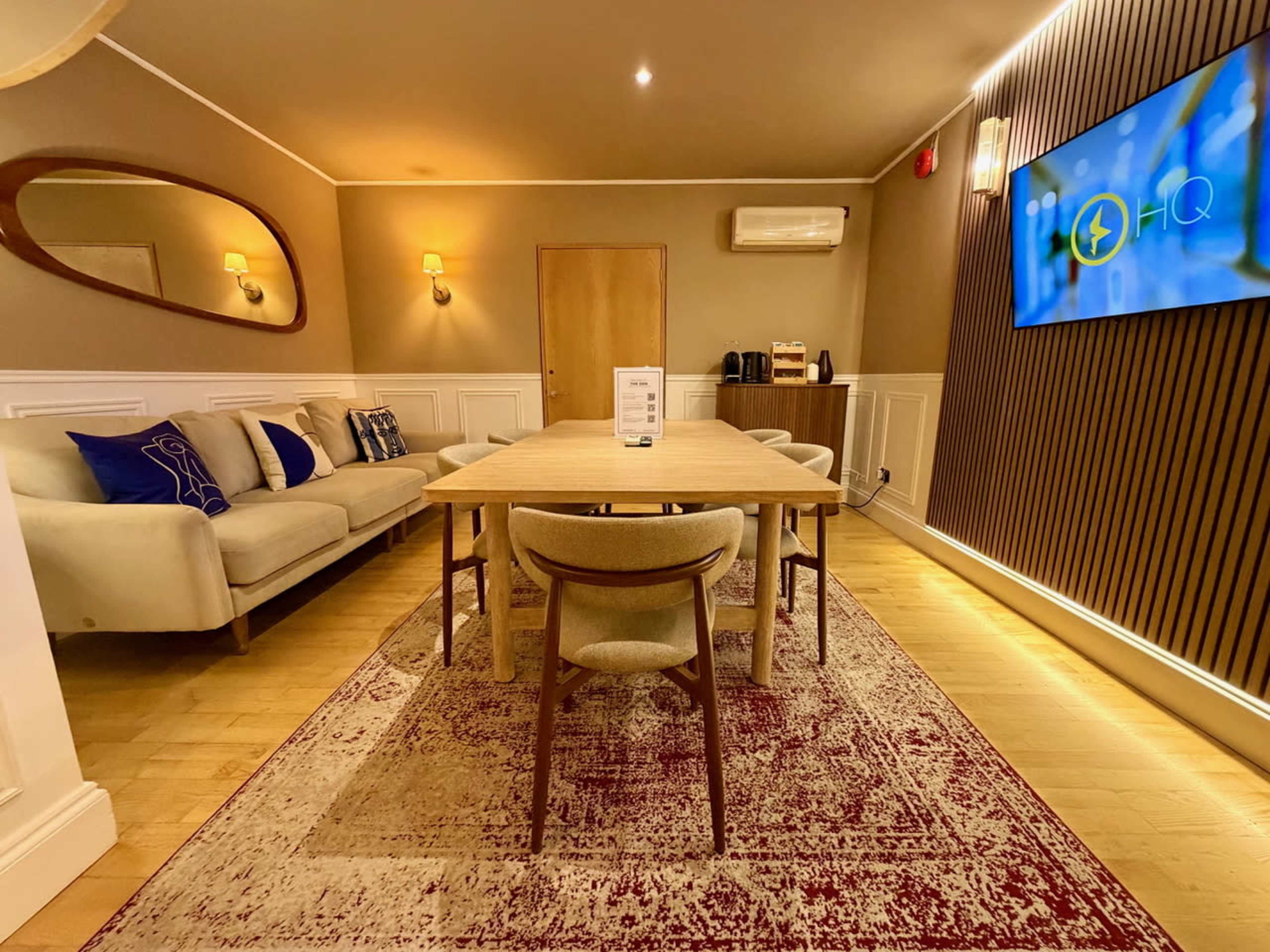 A modern meeting room features a long wooden table surrounded by chairs, a sofa, and an illuminated wall with a screen displaying a logo.