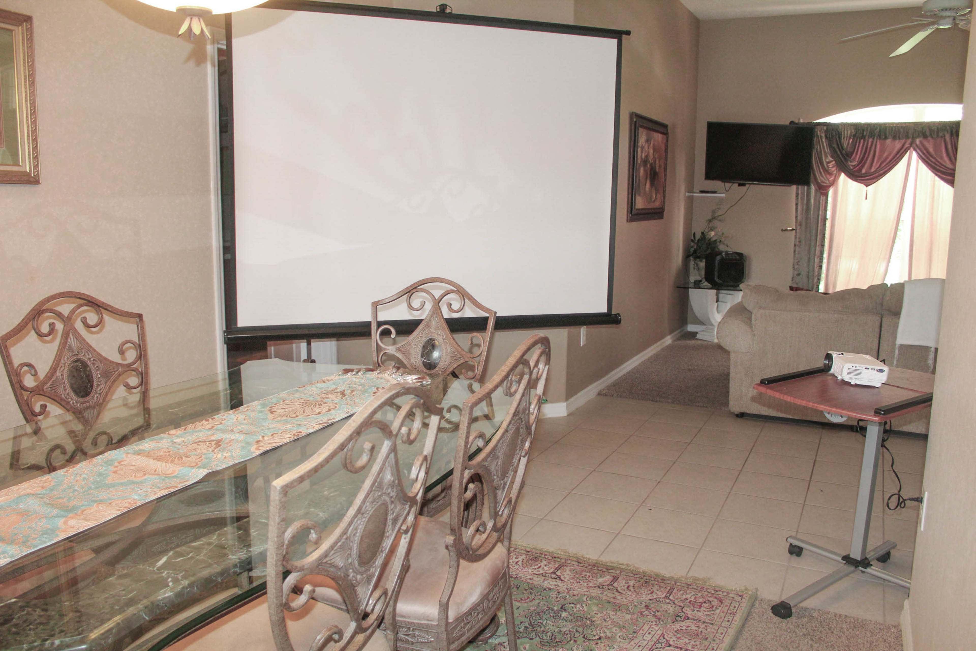 A dining area with a glass table and four chairs, a pull-down projector screen, and a small couch in a room with beige walls and carpet.