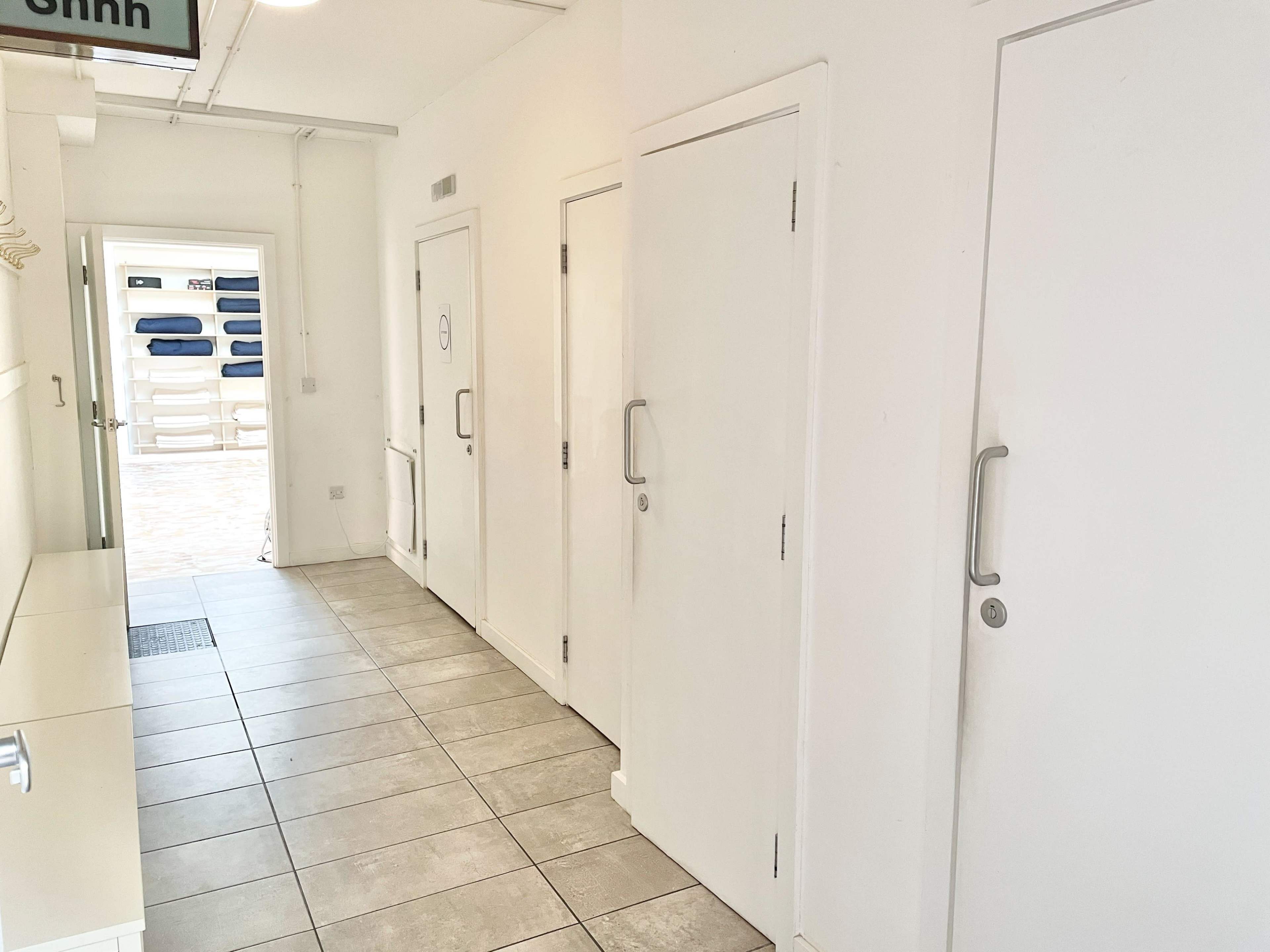 The image shows a hallway with white walls and multiple doors leading to separate rooms, with a glimpse of outdoor lounge chairs visible at the end.