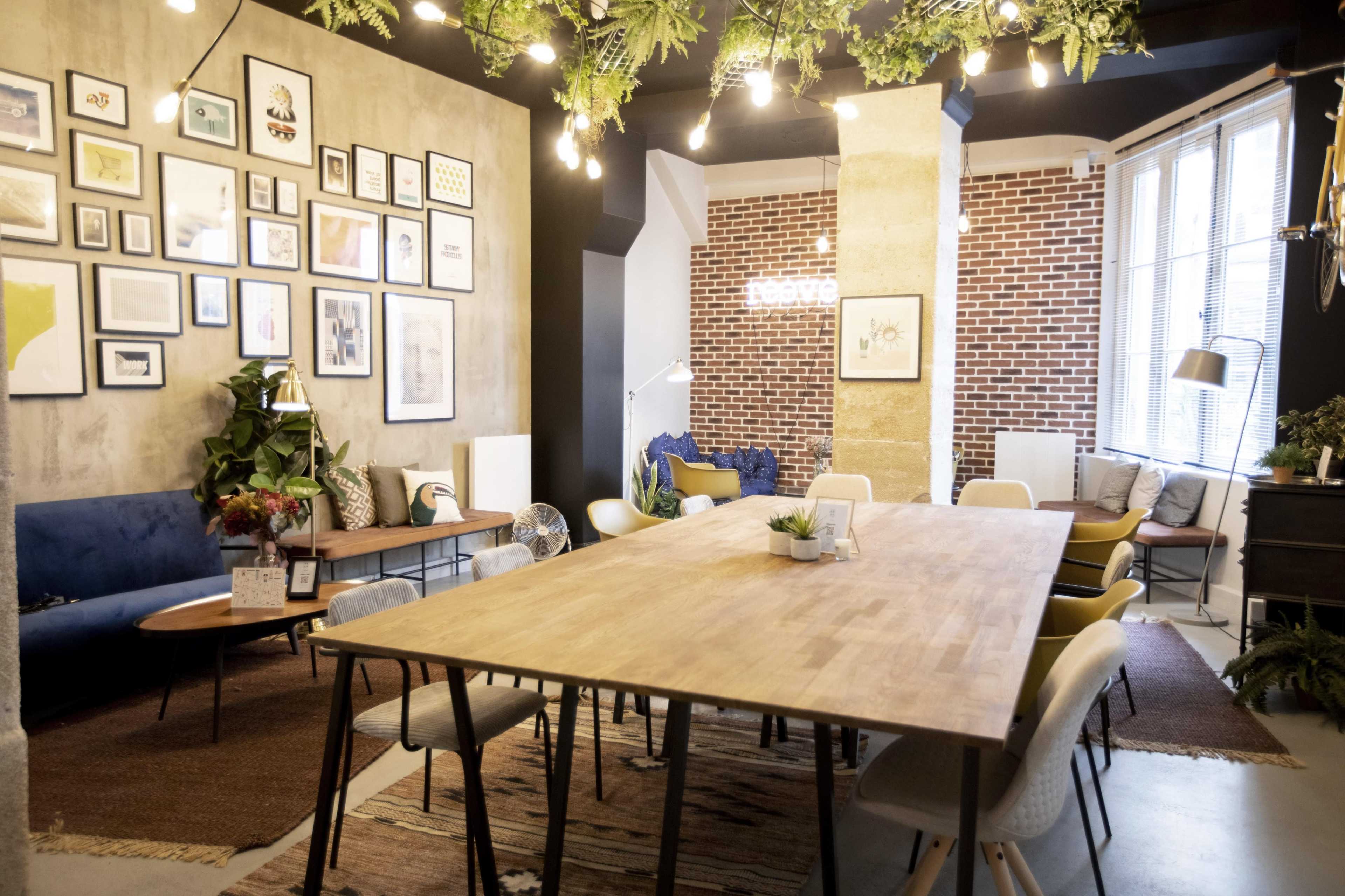 The image shows a modern meeting room featuring a long wooden table surrounded by chairs, framed artwork on the walls, and a mix of plants and decorative elements.