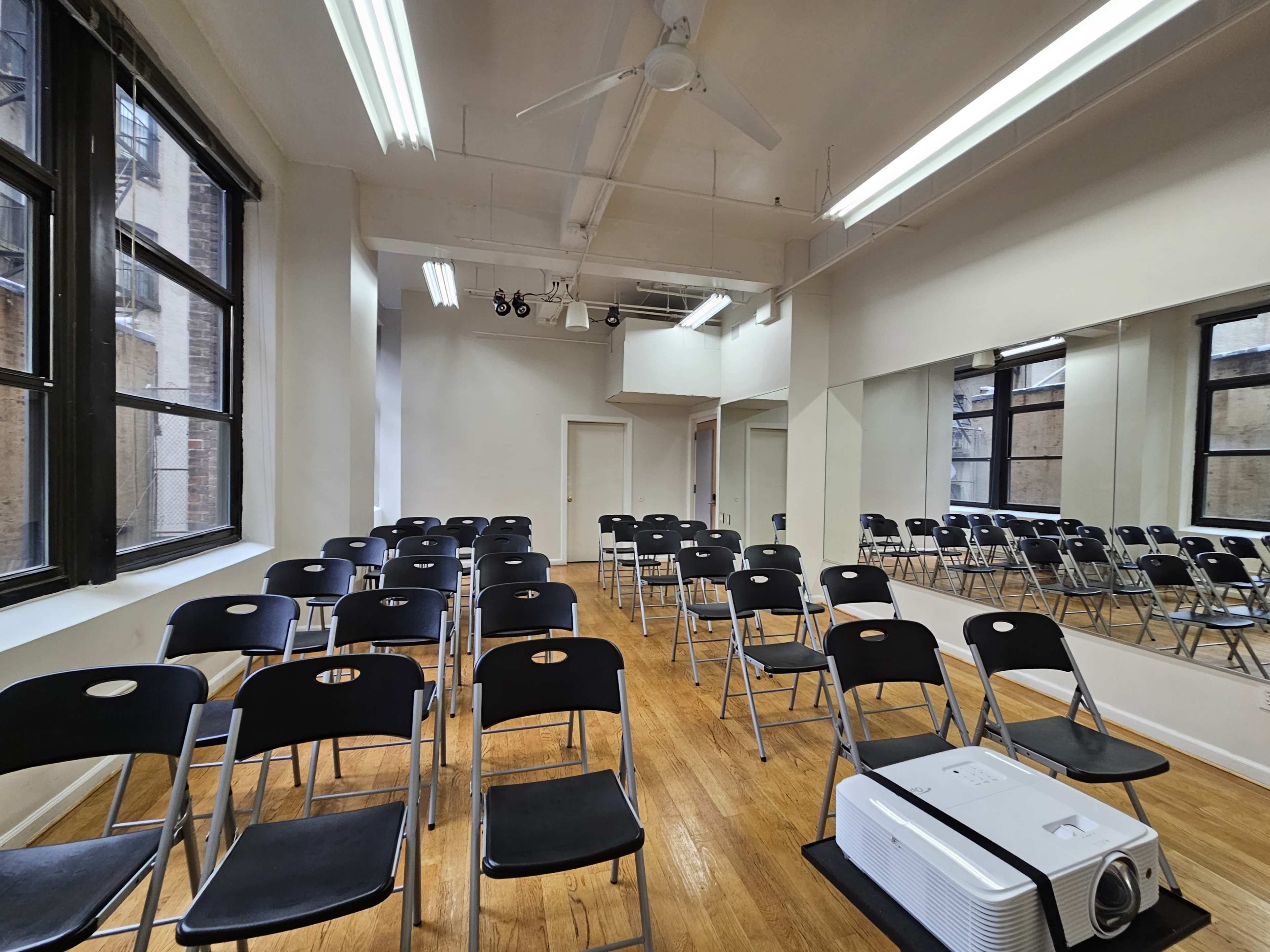 The image shows a spacious room with rows of black folding chairs facing a white projector placed on the wooden floor, and mirrors along one wall.