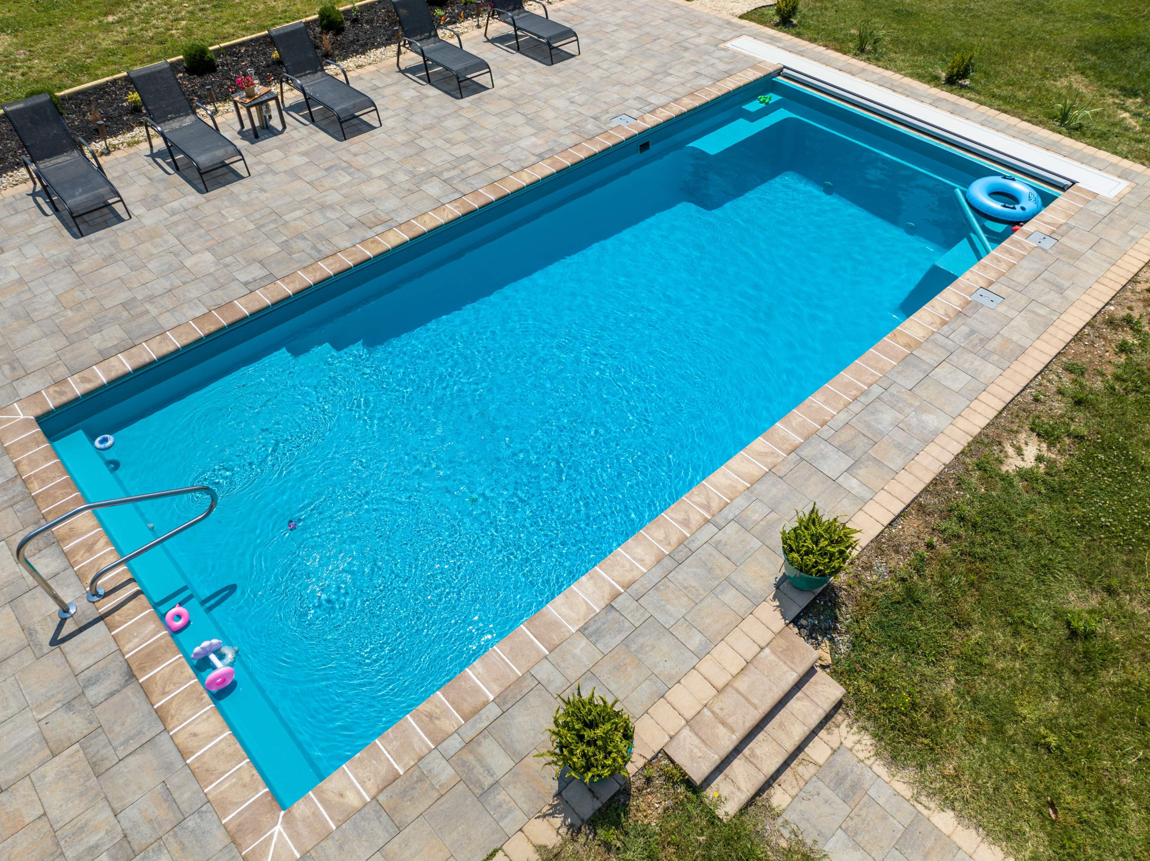 The image shows a swimming pool with a clear blue water surface, surrounded by a paved patio and several lounge chairs.