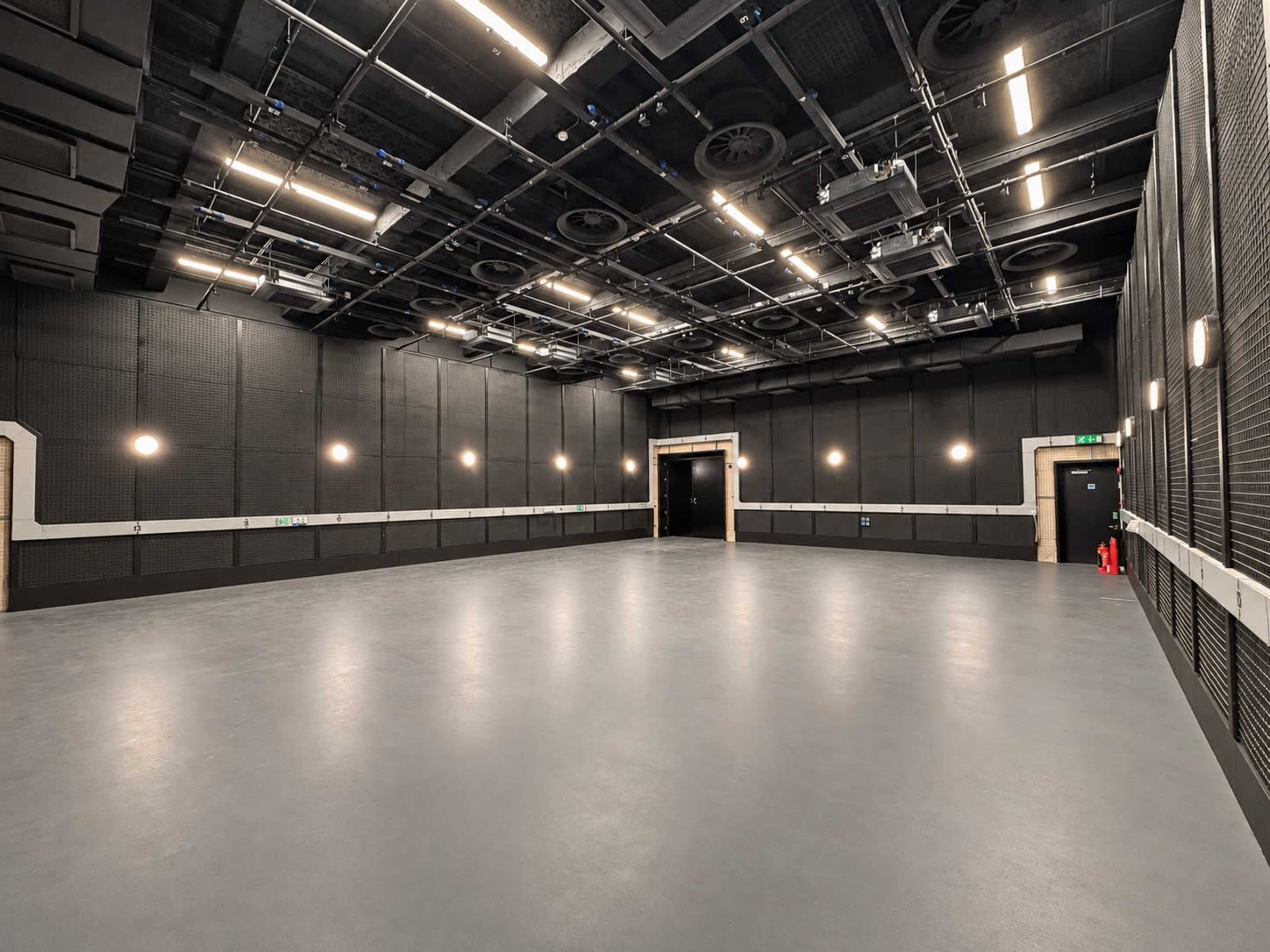 The image shows a large, empty studio space with black walls, a smooth gray floor, and overhead lighting fixtures.