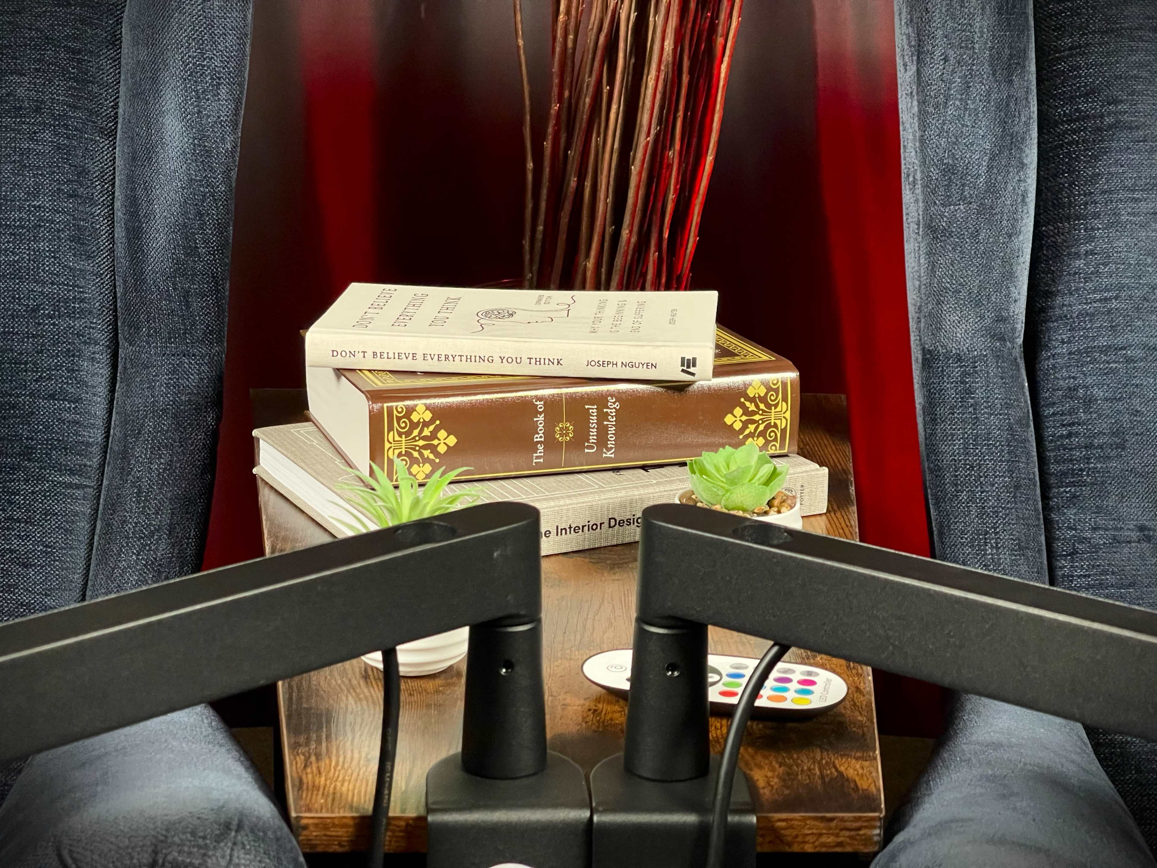 A cozy reading nook features two comfortable chairs, a stack of books, decorative plants, and a remote control on a wooden side table.