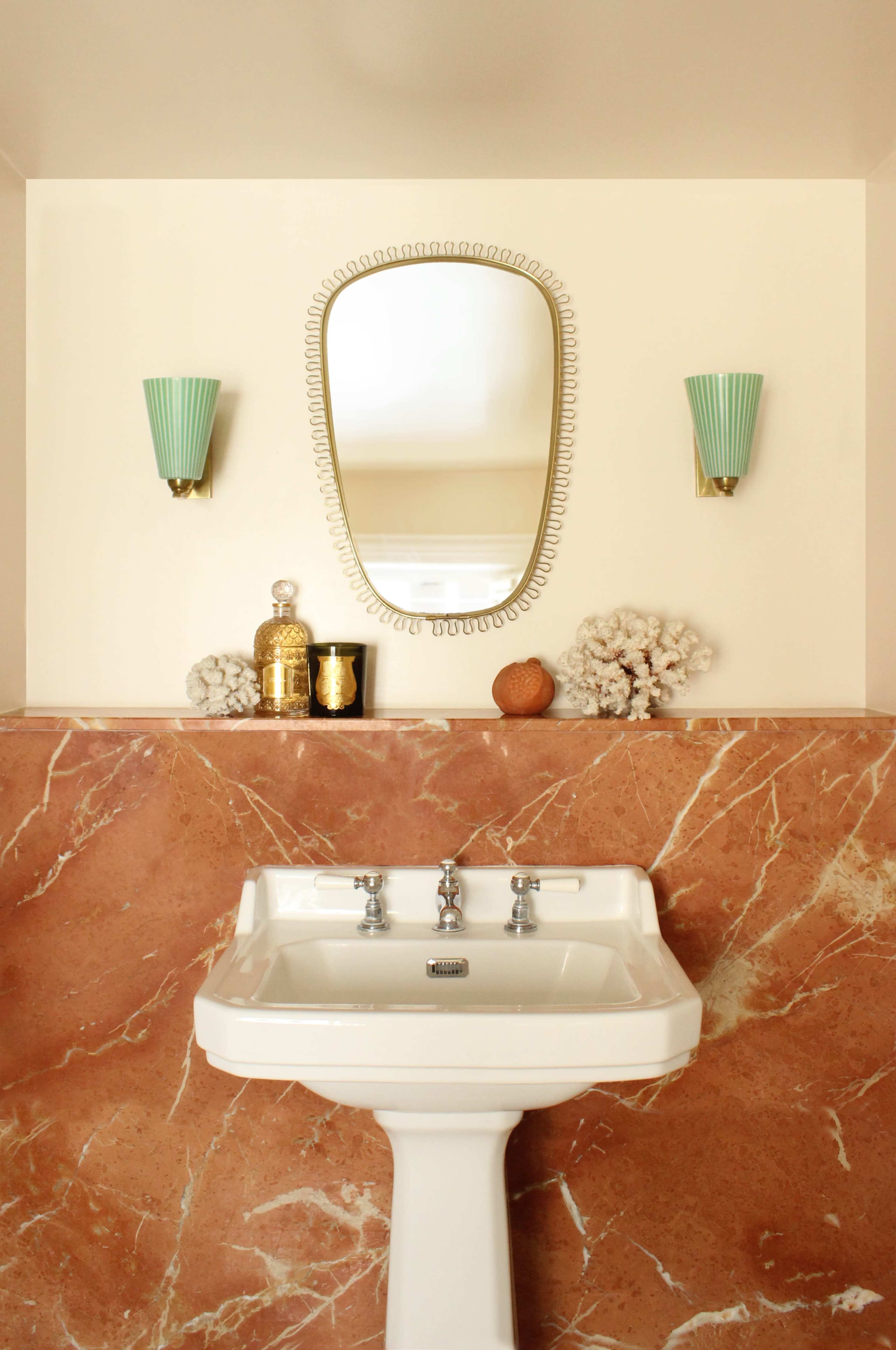 The image shows a bathroom with a white sink beneath a decorative mirror, flanked by green wall sconces, set against a pink marble backdrop.