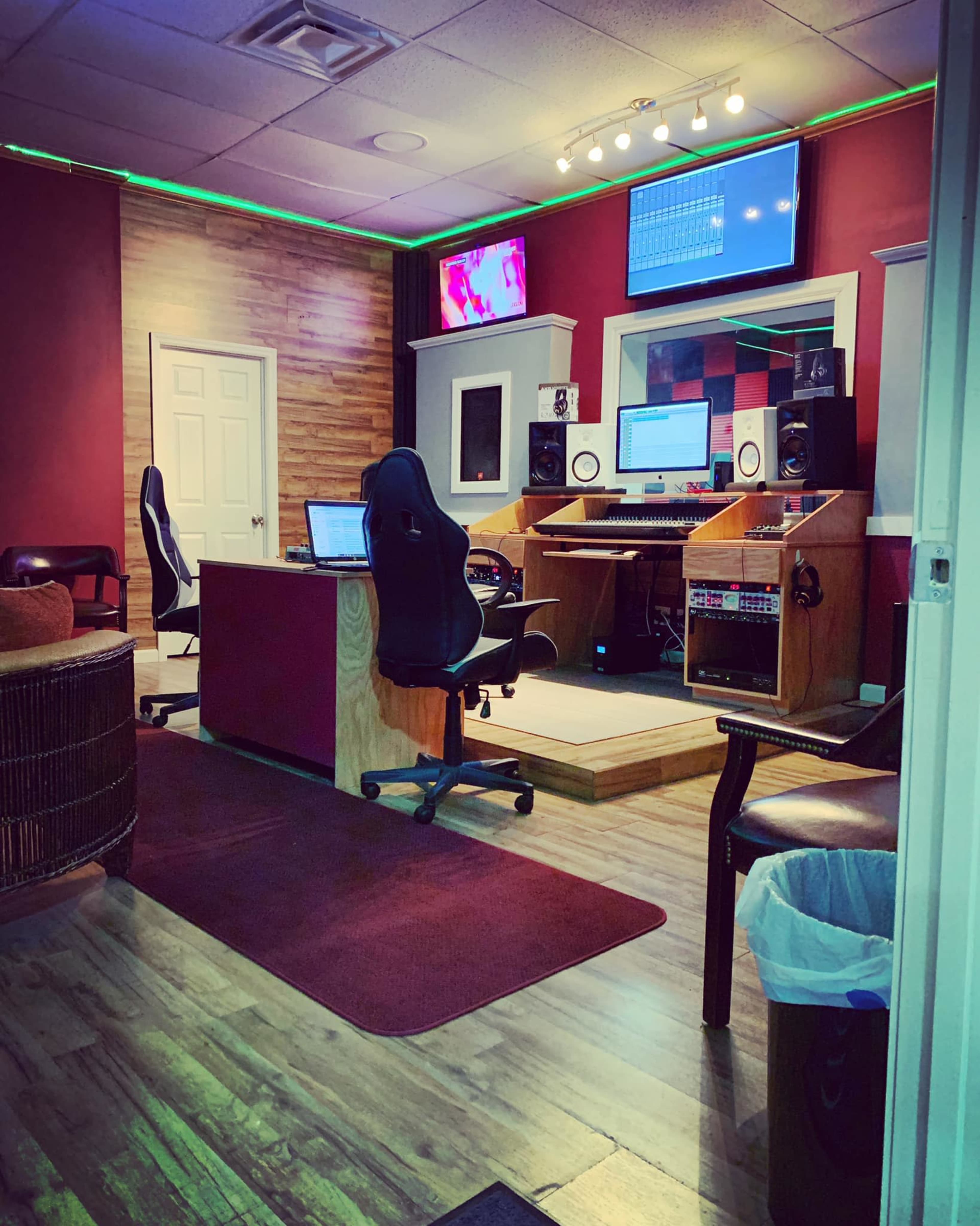 The image shows a recording studio with a control desk, monitors, and audio equipment, featuring wooden accents and a red wall.