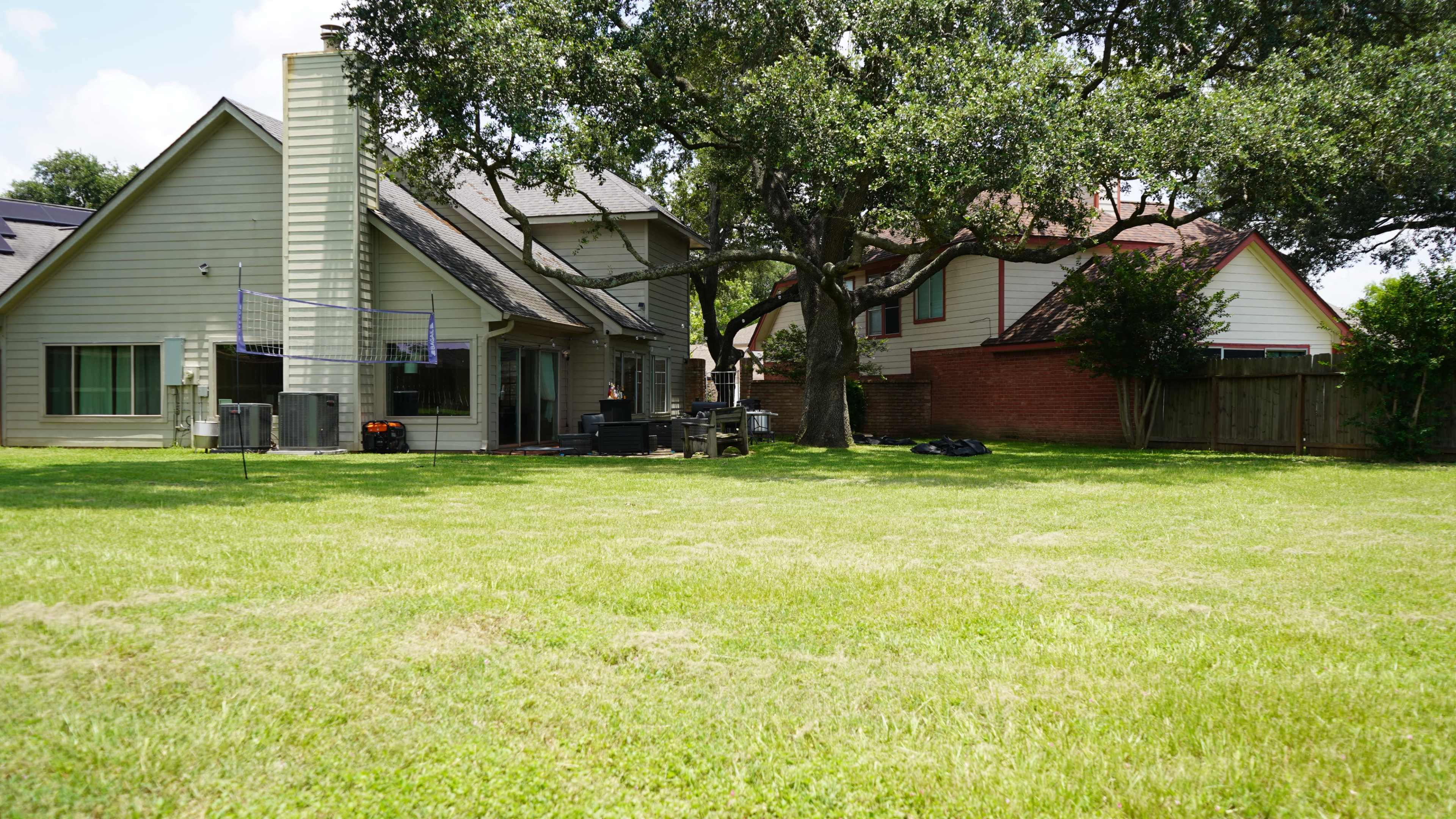 The image shows a backyard with a green lawn and several houses, including a two-story structure and a single-story building, with a large tree in the center.