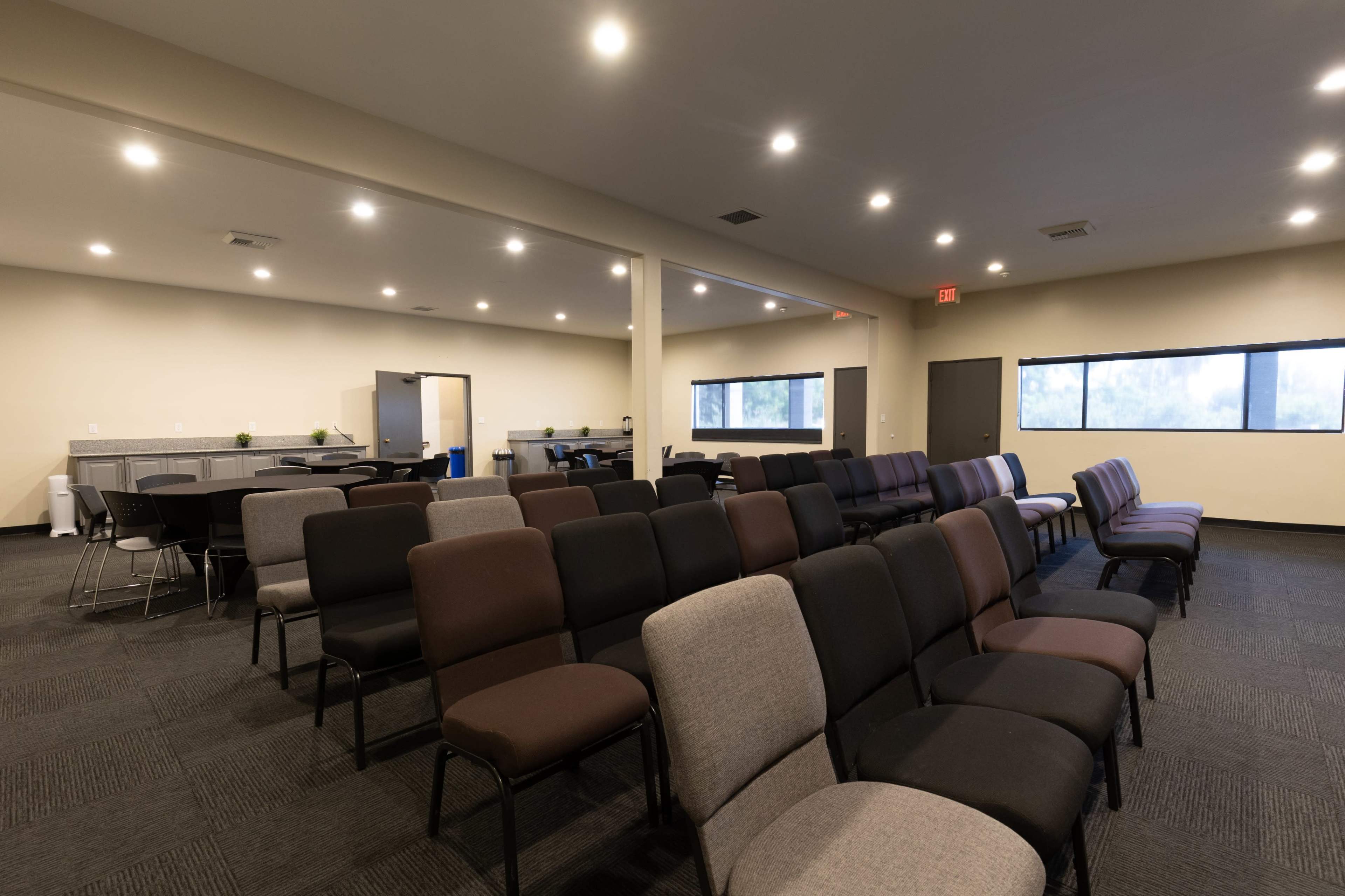 The image shows a spacious room arranged with rows of chairs, a kitchenette area, and a few tables near large windows.