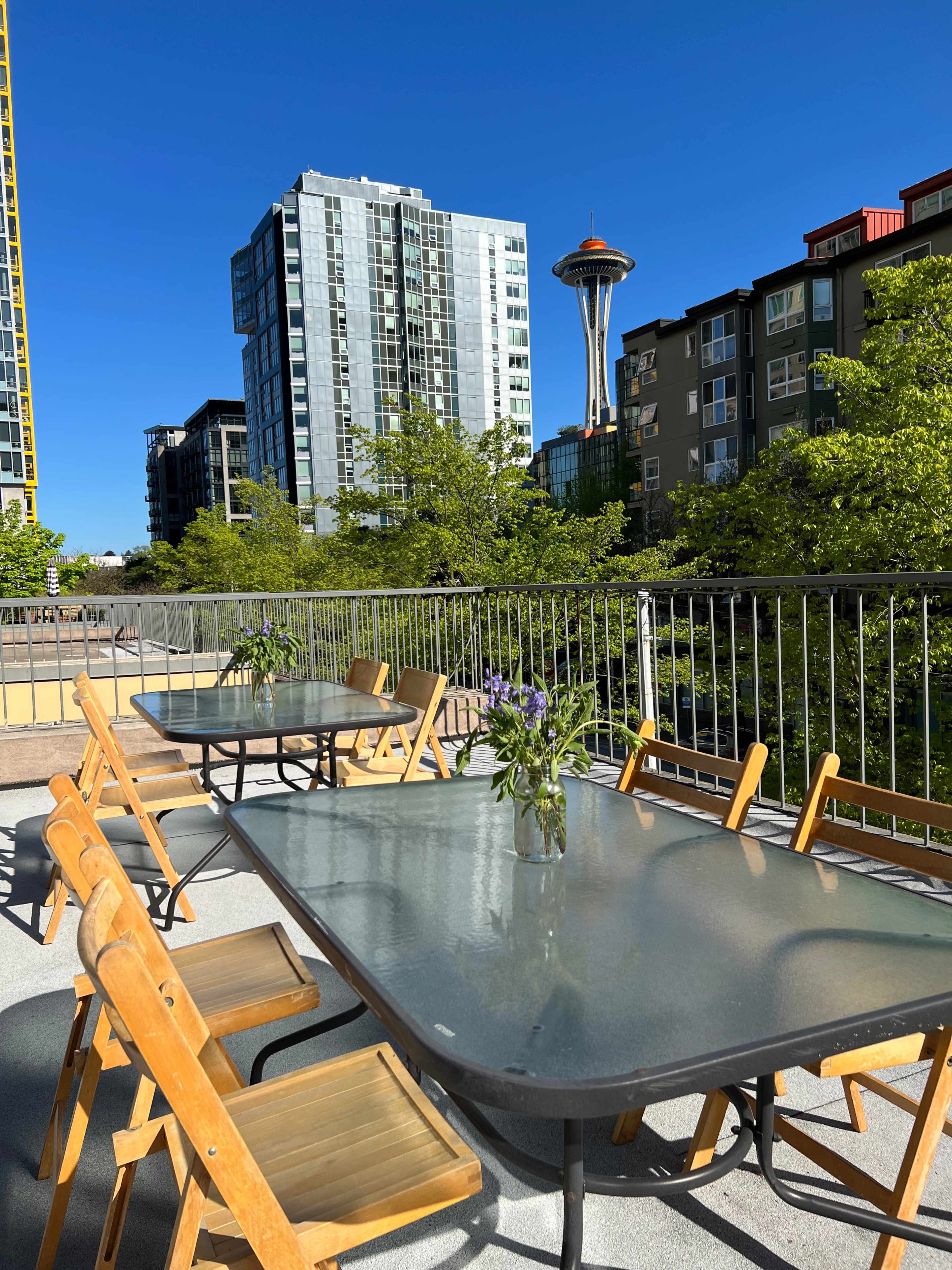 A rooftop deck features several wooden tables and chairs, with the Space Needle visible in the background among modern buildings and greenery.
