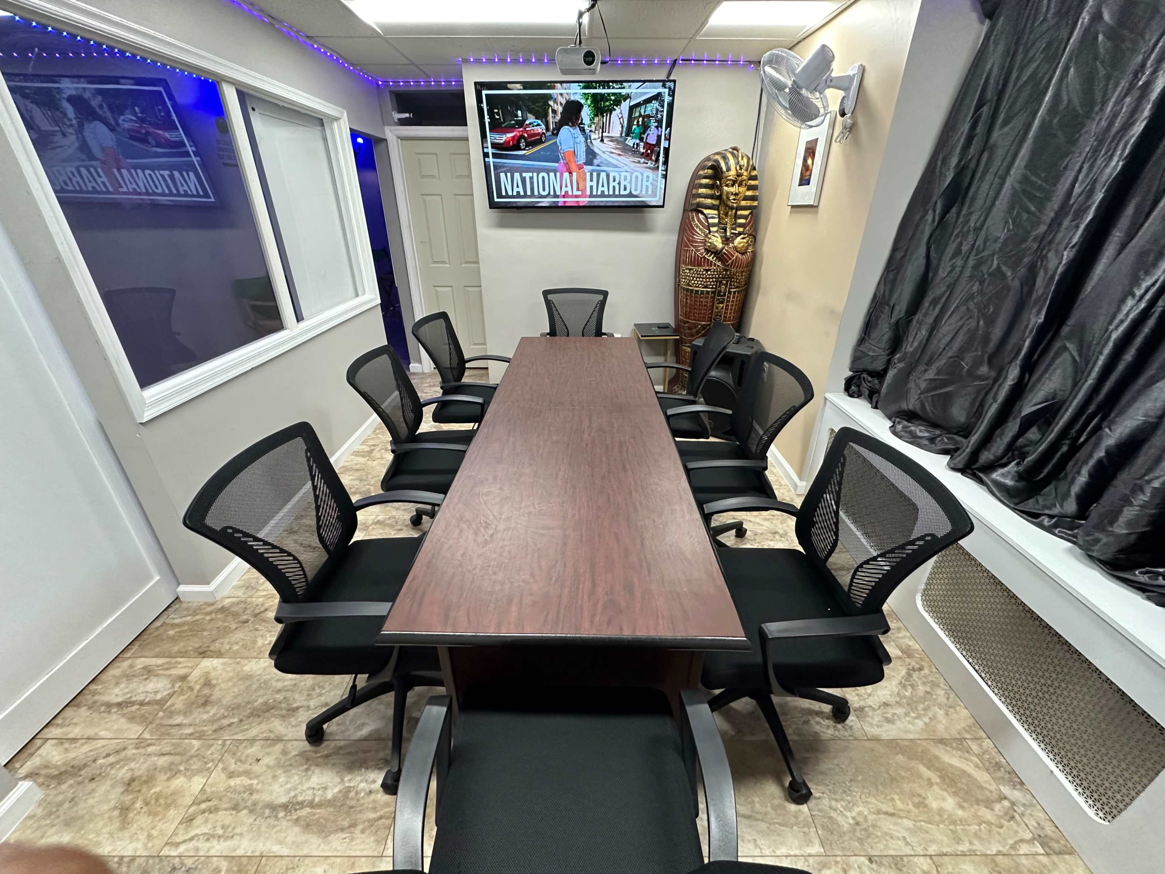 A conference room features a long wooden table surrounded by black mesh chairs, with a monitor displaying "NATIONAL HARBORS" on the wall.
