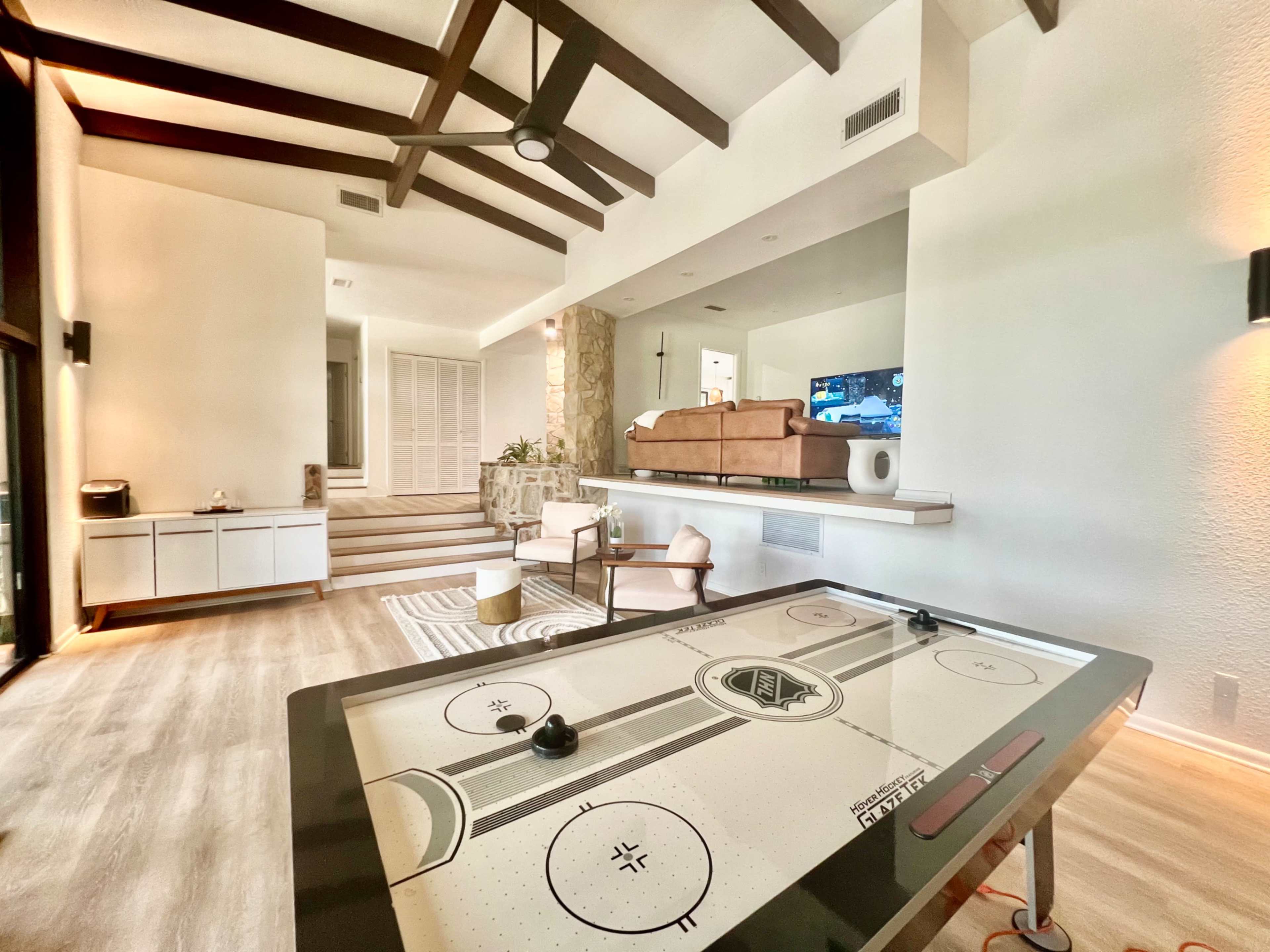The image shows a spacious living area featuring an air hockey table, a stone accent wall, and a sectional sofa in front of a television.