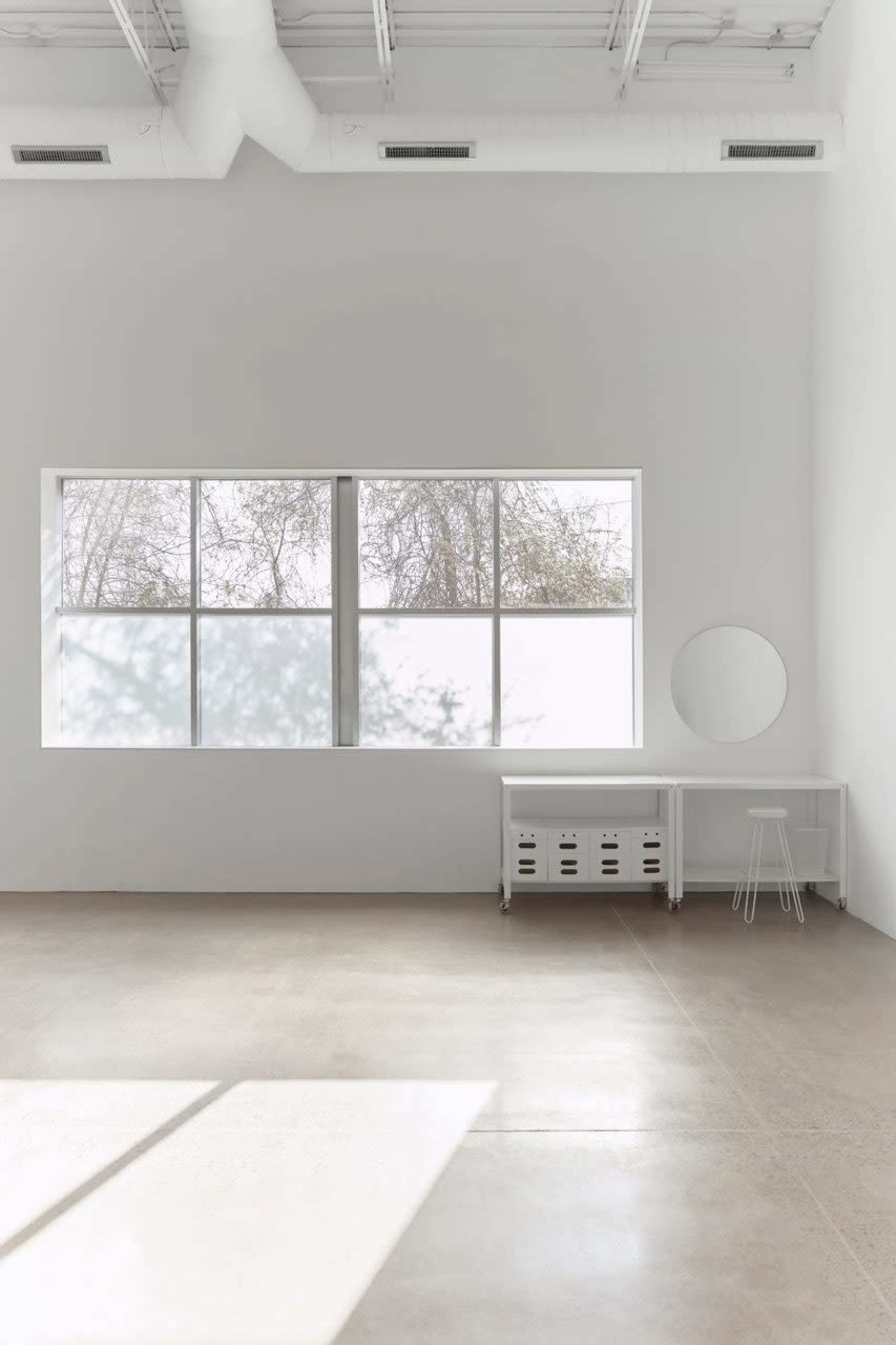 The image shows a spacious, minimalistic room with a large window, a white circular mirror, a small table, and a stool.