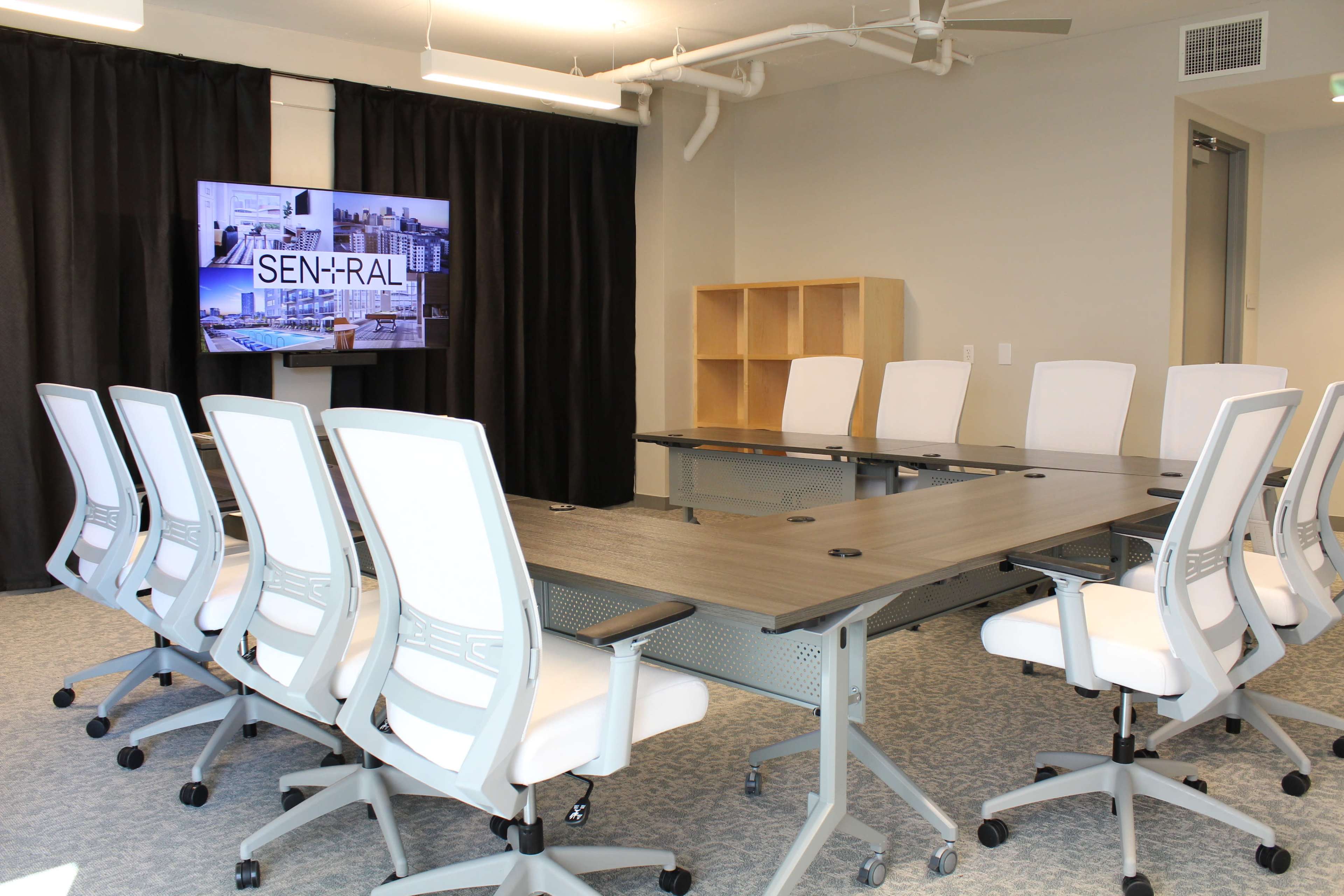 The image shows a modern conference room equipped with a long table, white ergonomic chairs, and a screen displaying the word "SEN+RAL" on a black curtain backdrop.