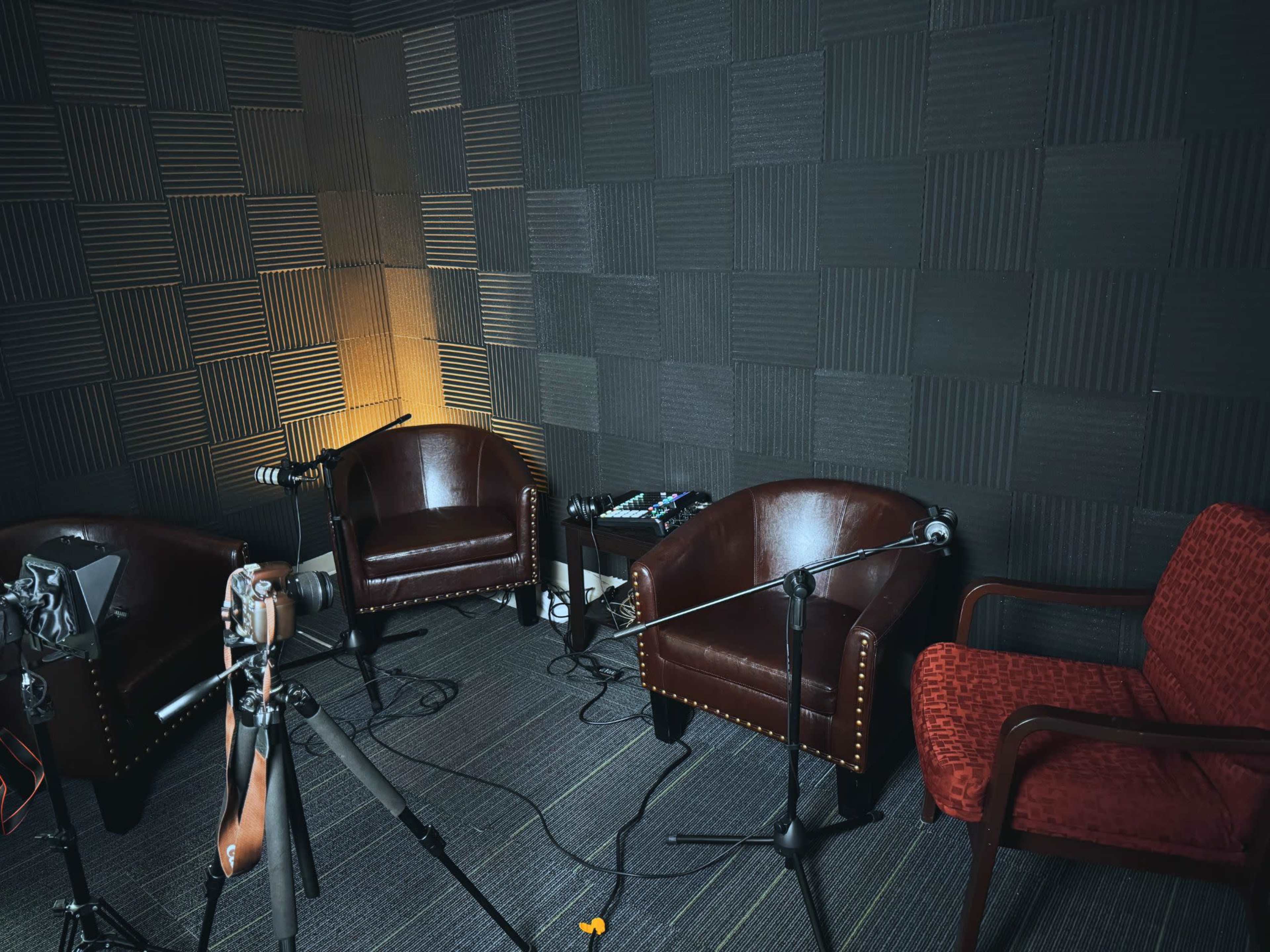 The image shows a recording studio with two brown leather chairs, a red fabric chair, and microphones set up on stands beside a tripod and audio equipment.