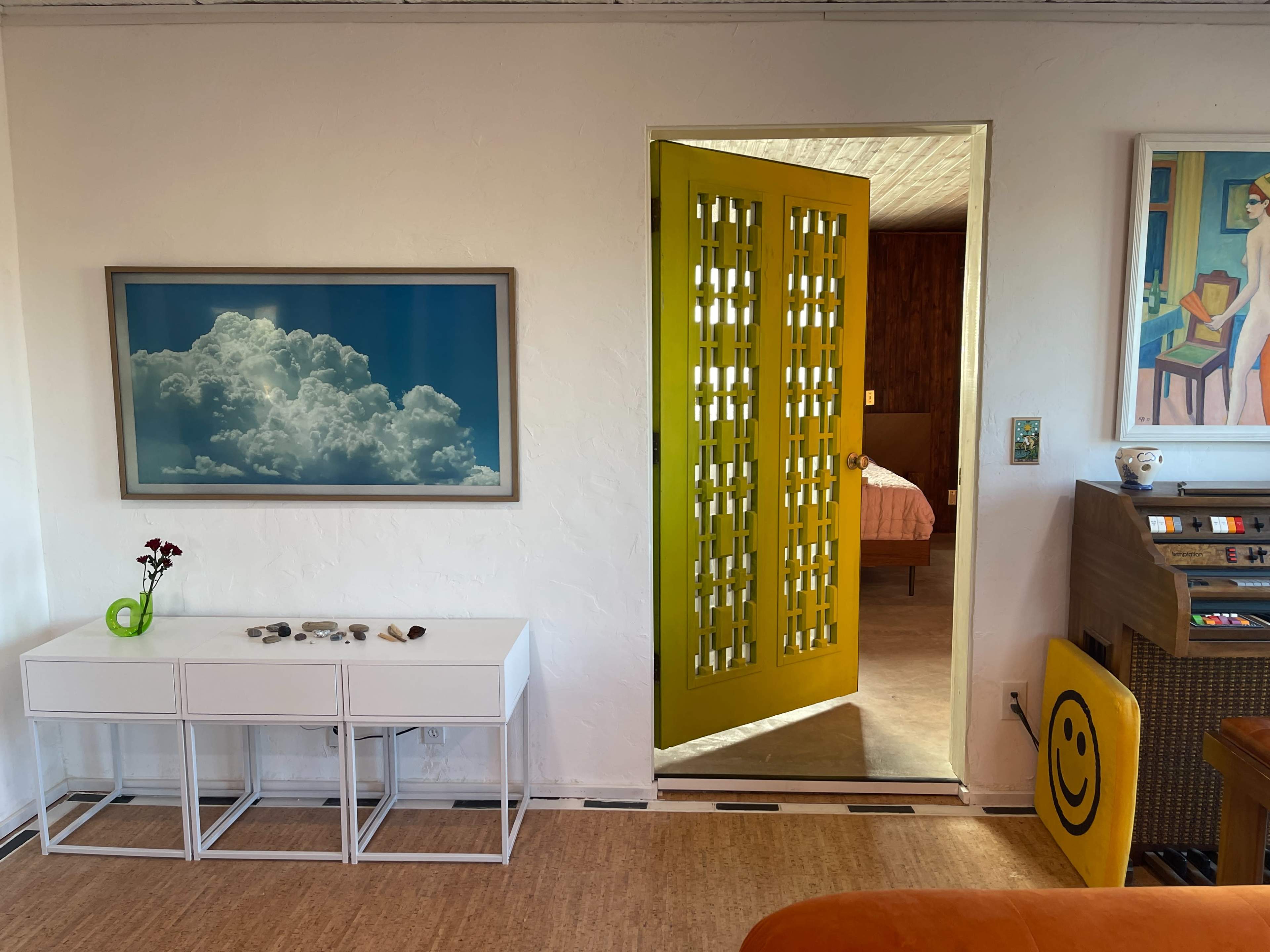The image shows a brightly colored room with a white side table, a cloud painting on the wall, and a yellow door leading to another space.