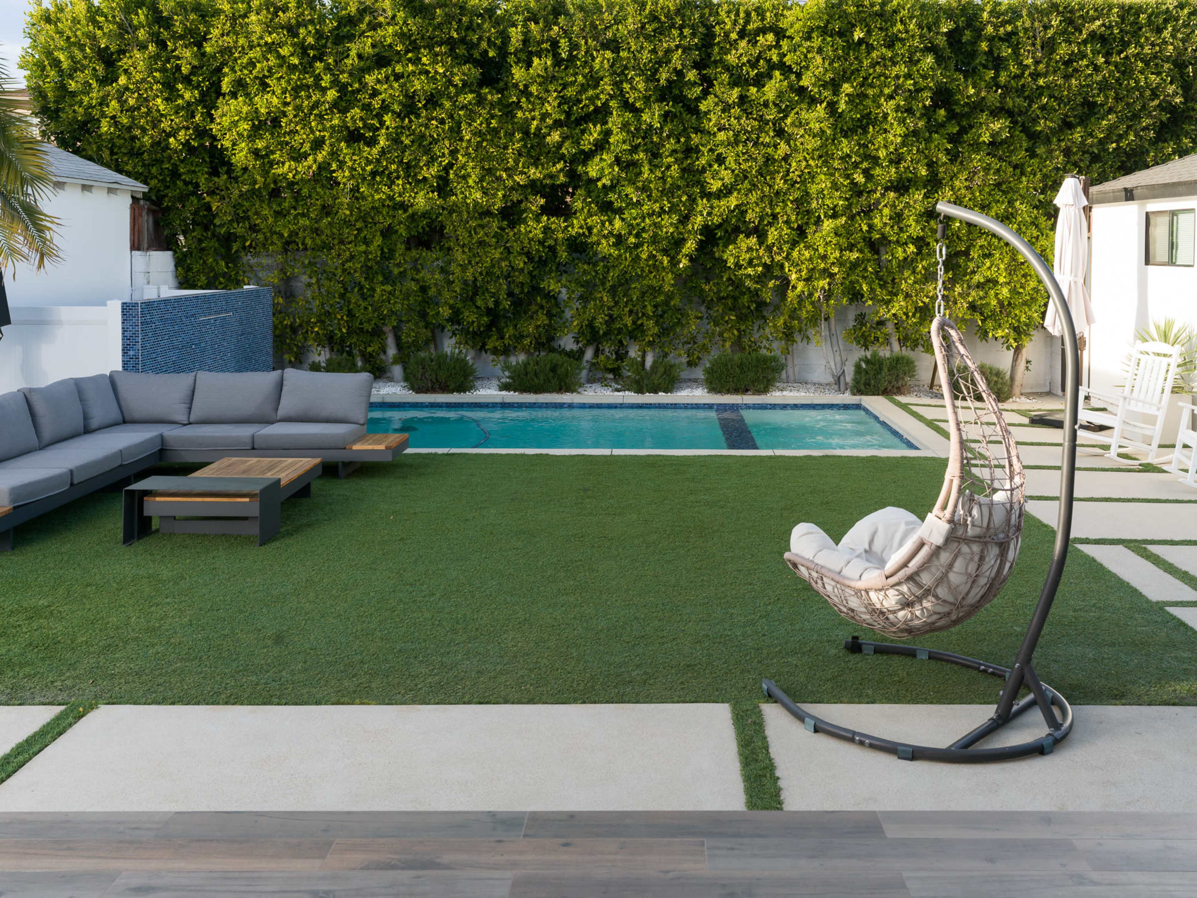 A modern backyard featuring a swimming pool, a large sectional sofa, and a hanging chair on a grassy area.