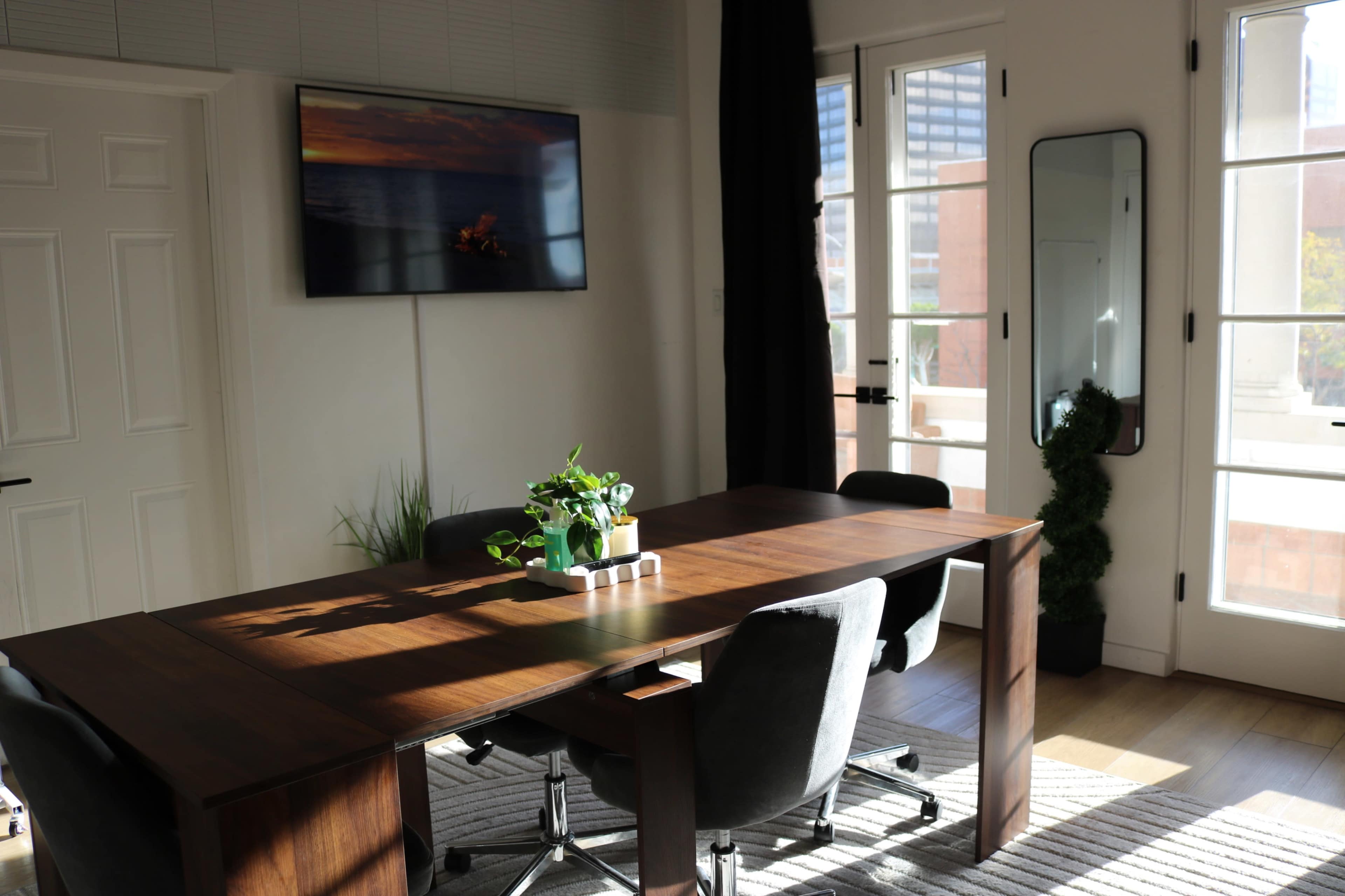 A modern conference room features a wooden table surrounded by black chairs, with a television mounted on the wall and large windows letting in natural light.