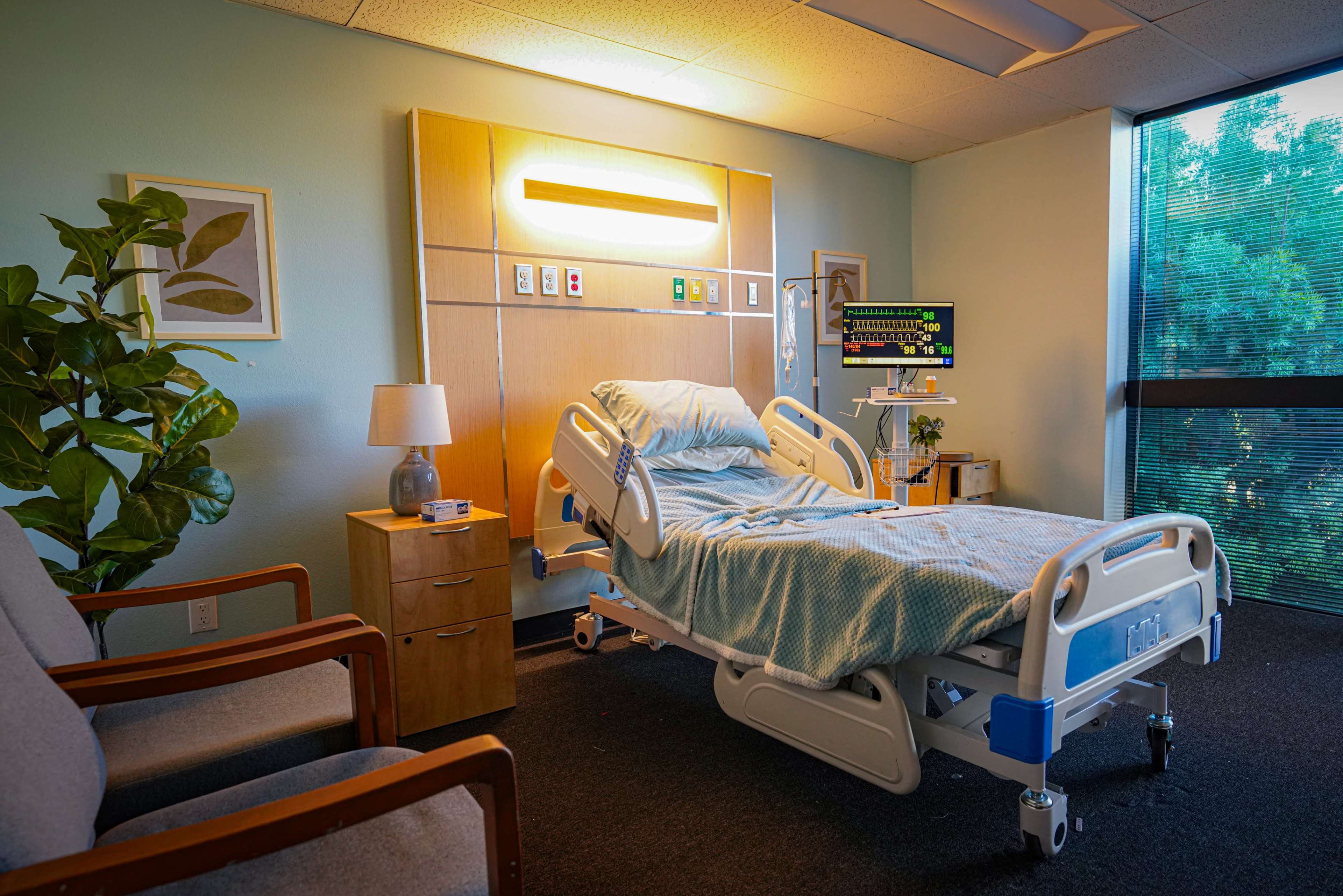 The image shows a hospital room featuring a bed with medical equipment, a side table, a lamp, and two chairs by a window.