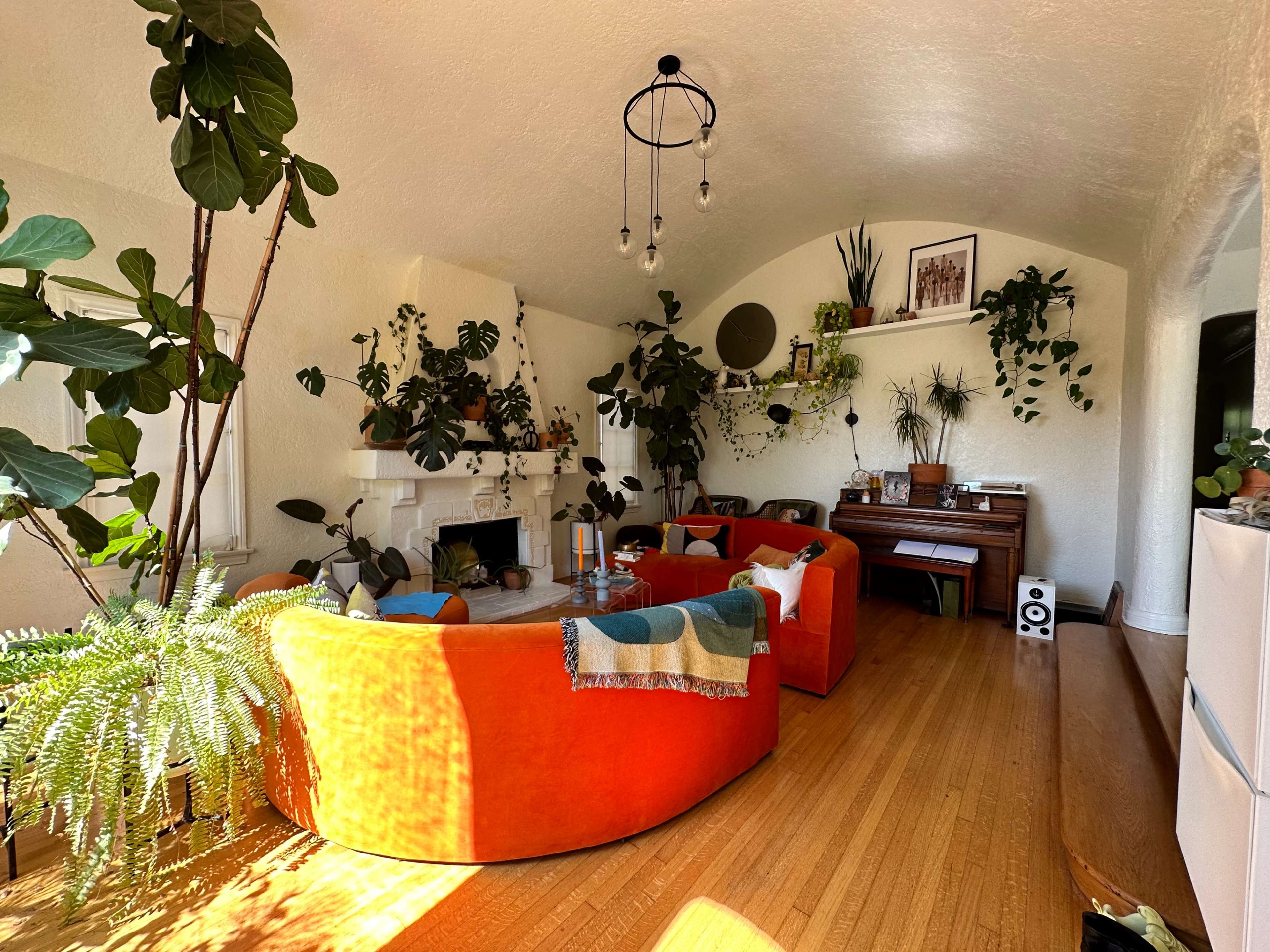 The image shows a cozy living room featuring a circular orange sofa, a wooden piano, and various indoor plants arranged around a fireplace.