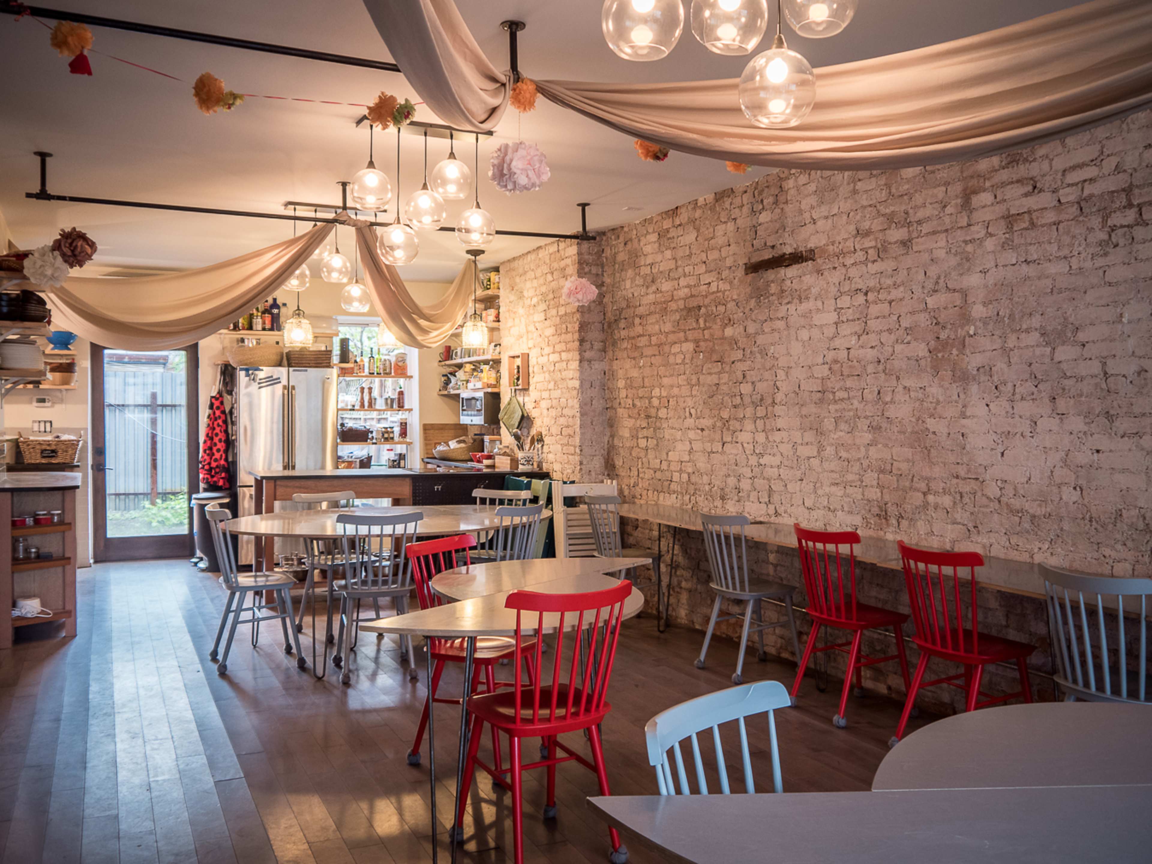 A cozy cafe interior featuring brick walls, wooden floors, and tables surrounded by colorful chairs under decorative lighting.