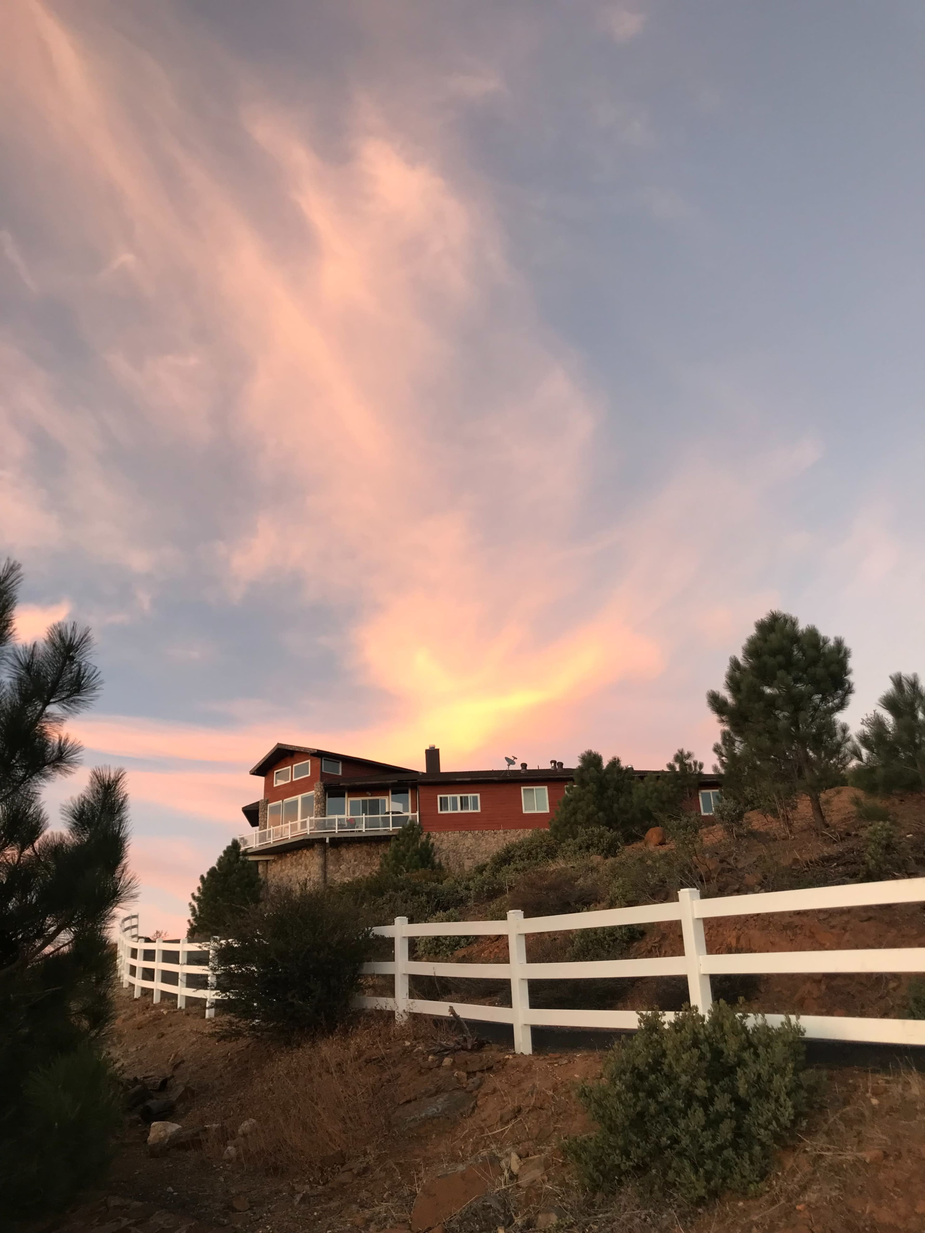 A house with a deck is situated on a hillside against a colorful sky at sunset, surrounded by trees and a white fence.