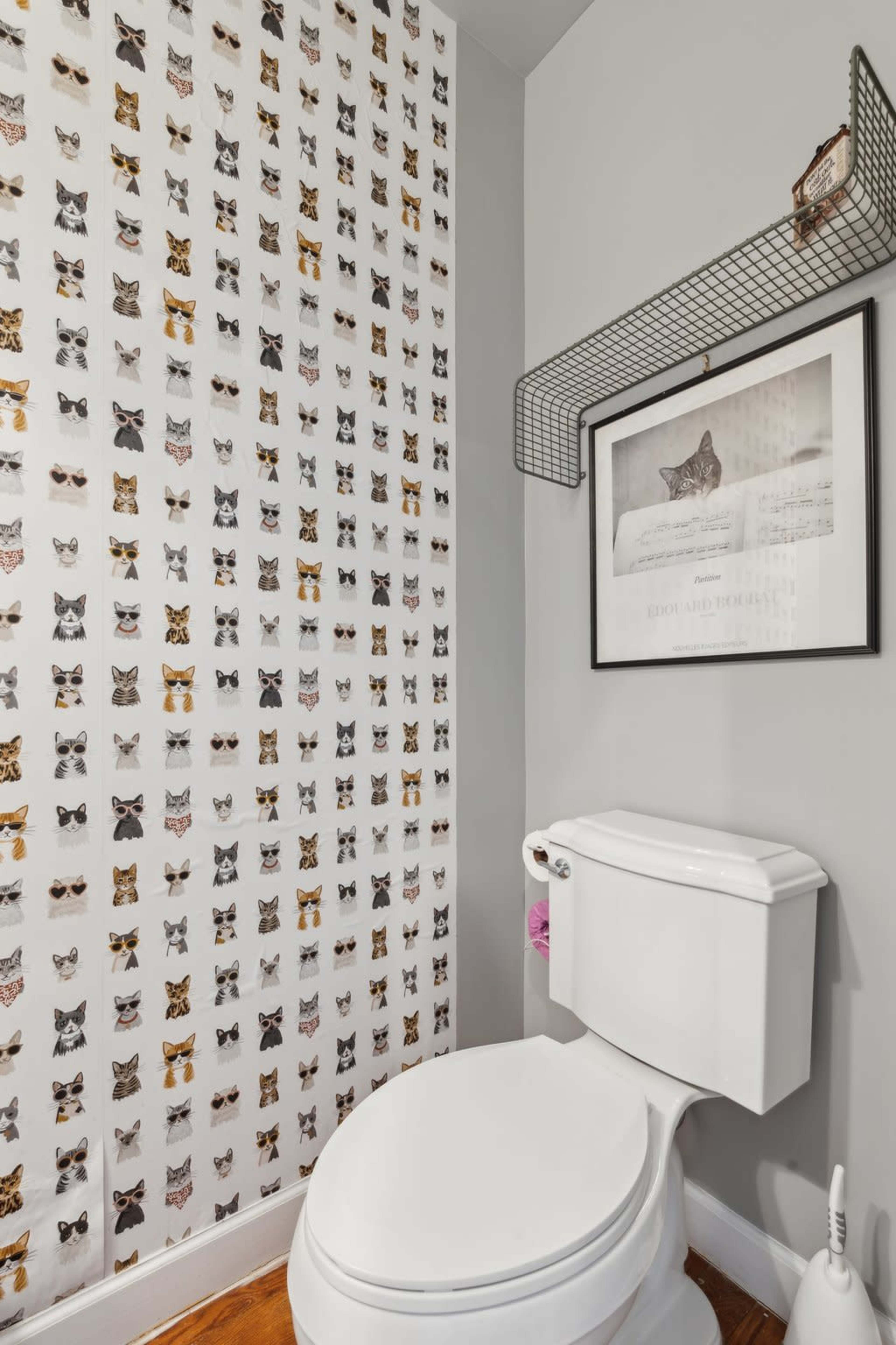 A bathroom features a white toilet and a wall decorated with a patterned wallpaper of cats in various poses, alongside a framed picture of a cat.