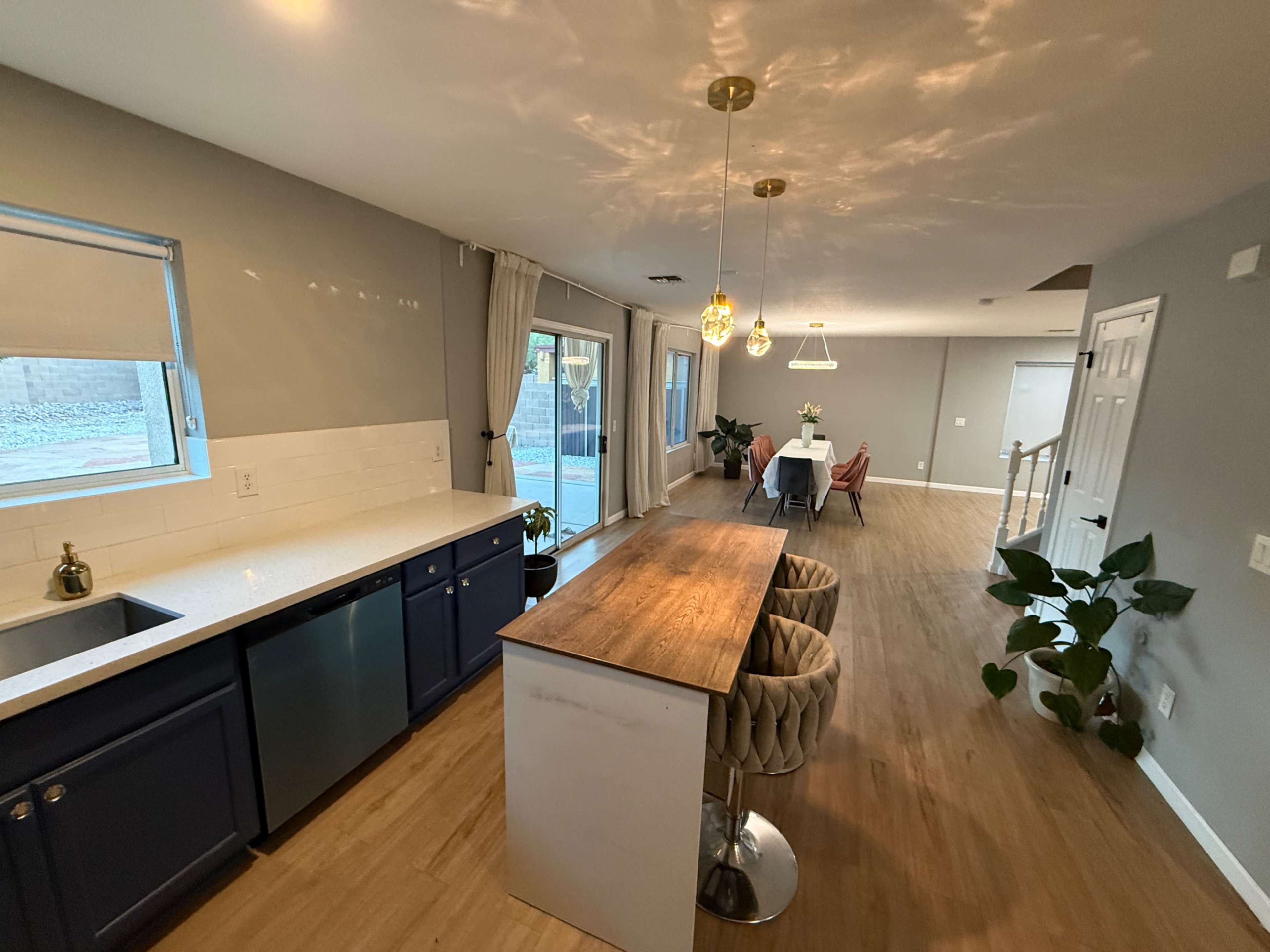 The image shows a modern kitchen and dining area featuring a large island with bar stools, a window with blinds, and a dining table in a well-lit space.