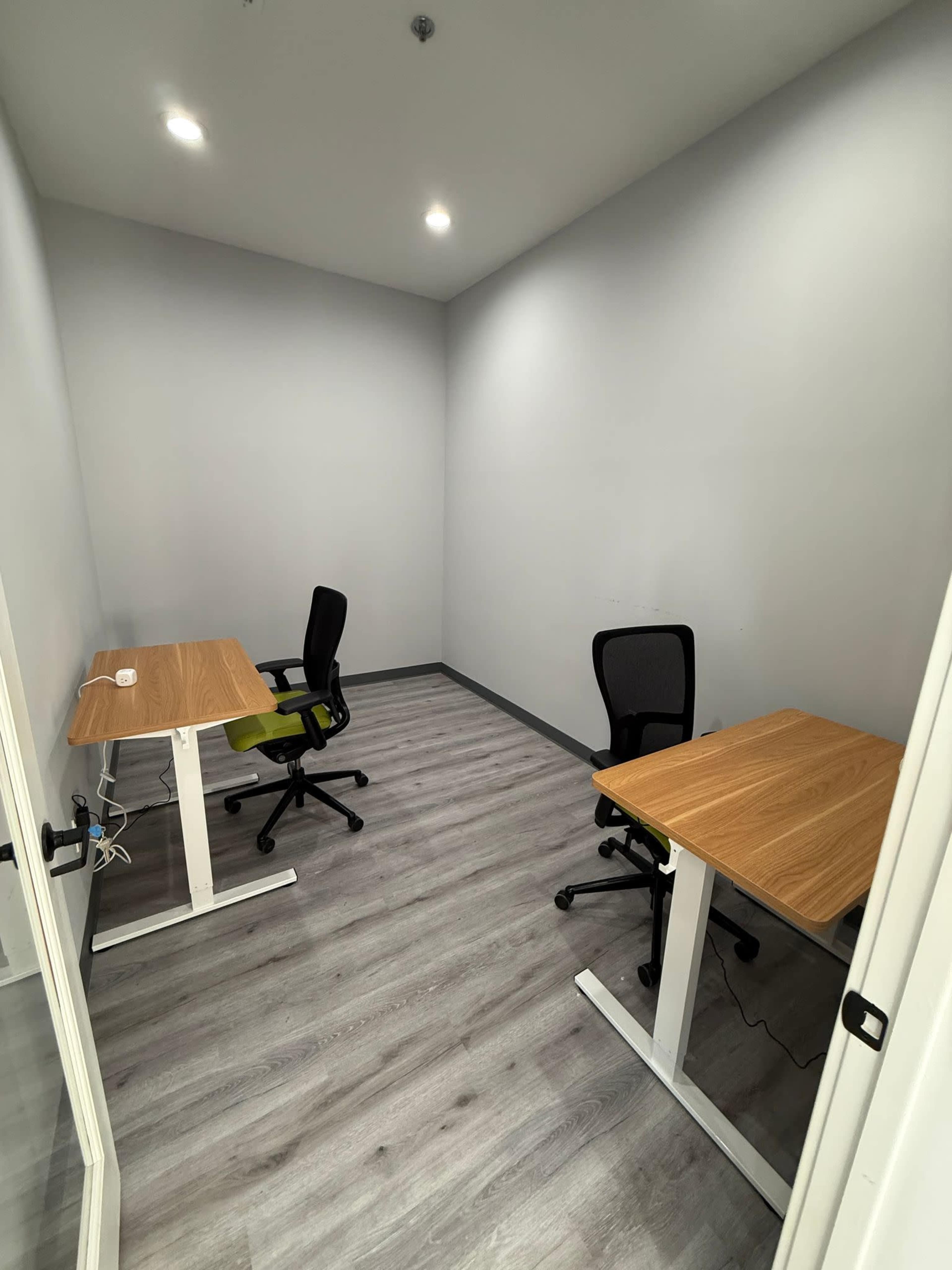 The image shows a small office space featuring two wooden desks with chairs, set against a neutral gray wall and laminate flooring.