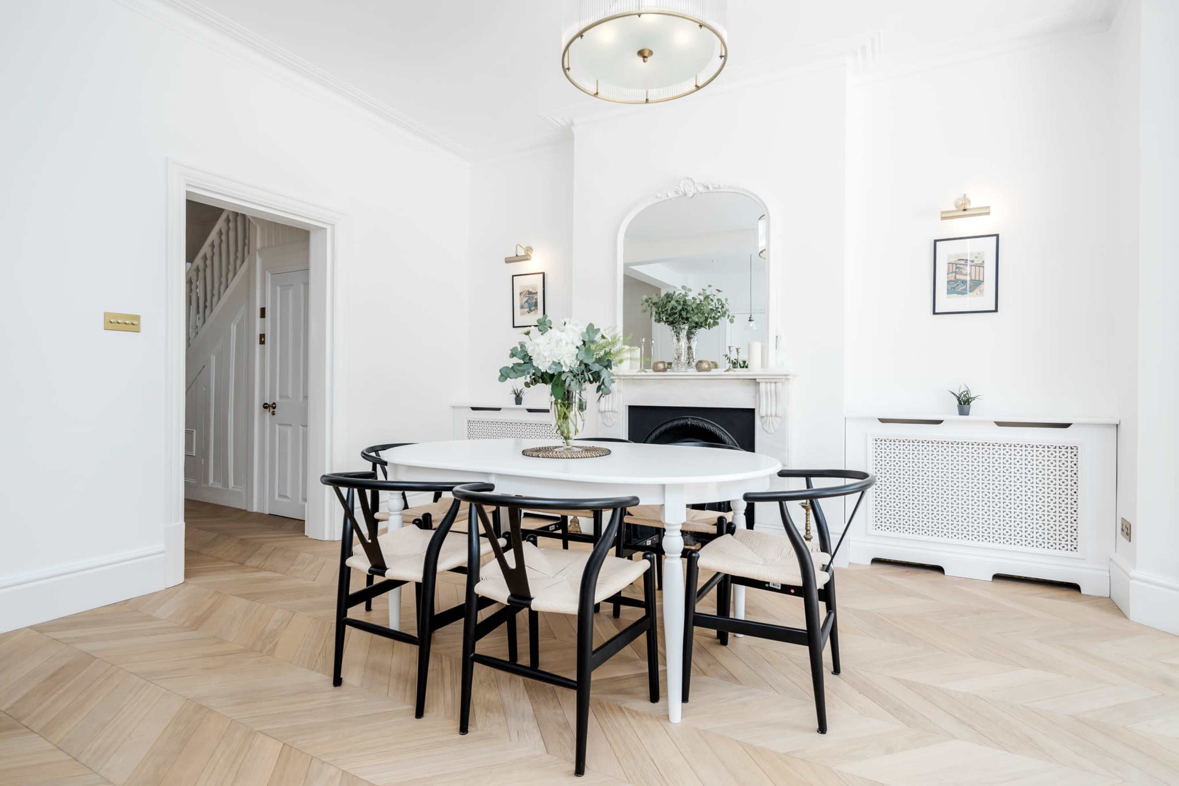 A modern dining room features a round white table surrounded by black chairs, with a fireplace and floral decor in the background.