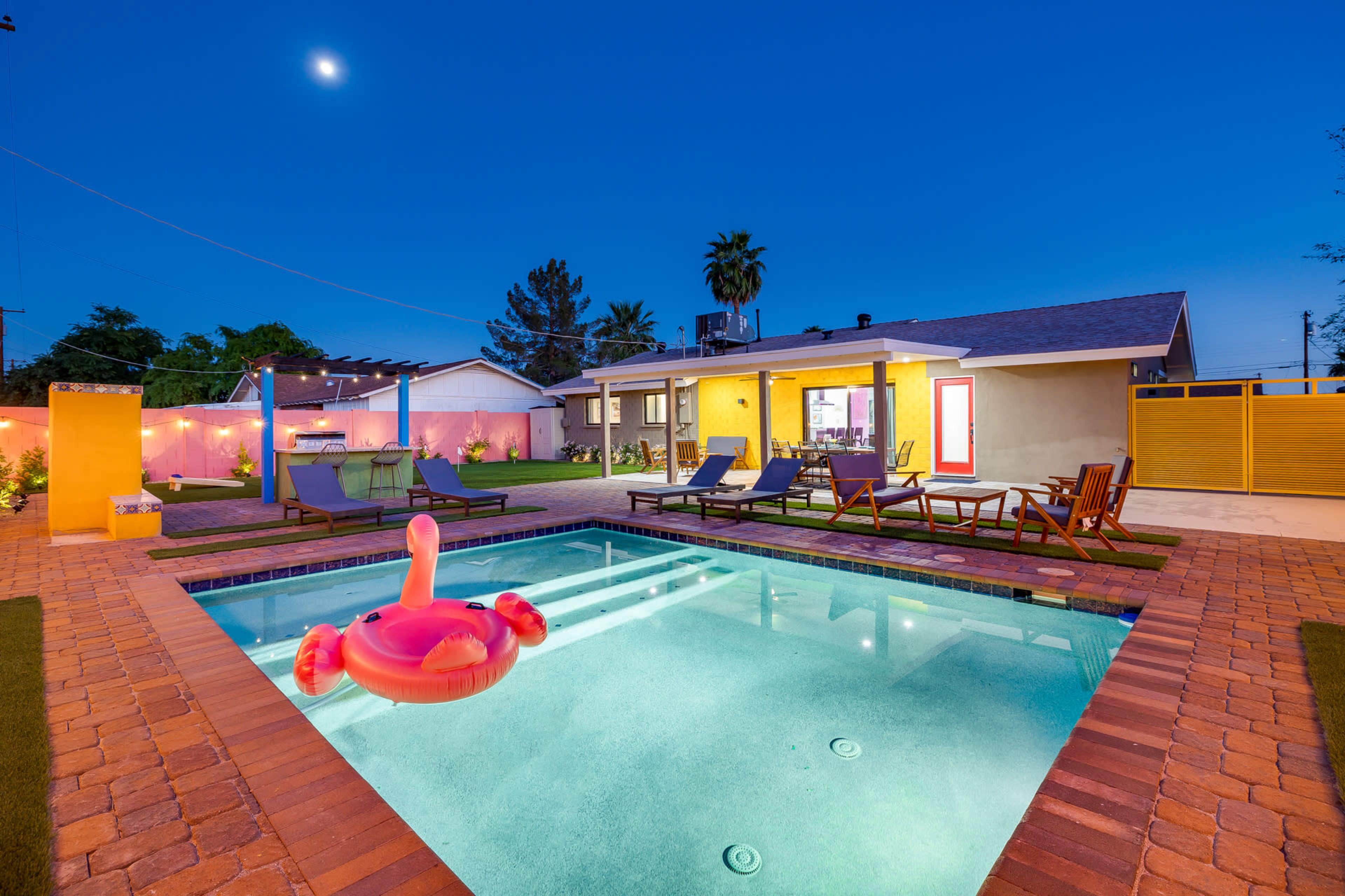 A swimming pool with a pink flamingo float is surrounded by lounge chairs and a well-lit backyard at dusk.