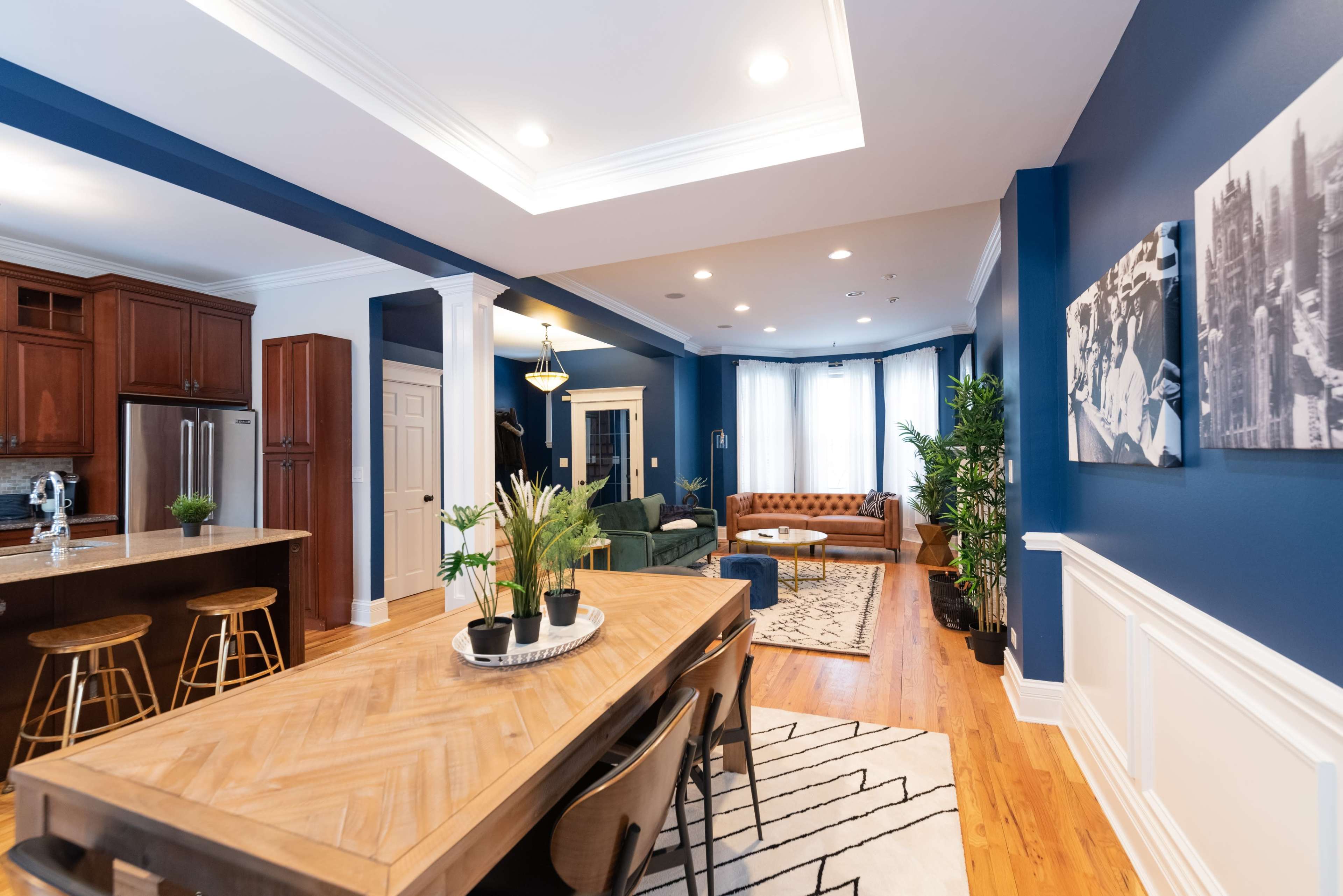 A spacious living and dining area features a wooden dining table, modern bar stools, a green sofa, and brown leather seating, with blue walls and large windows allowing natural light.