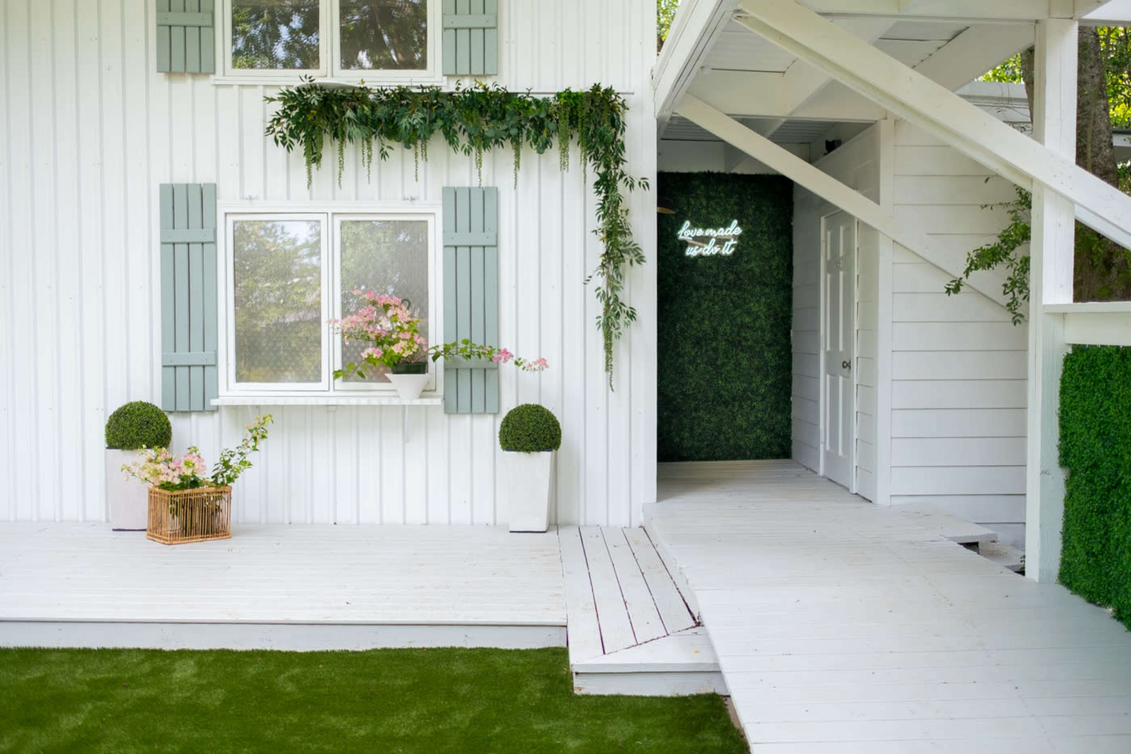 A white wooden house with green shutters, a flower-filled window box, and a porch leading to a door framed by a wall of greenery.