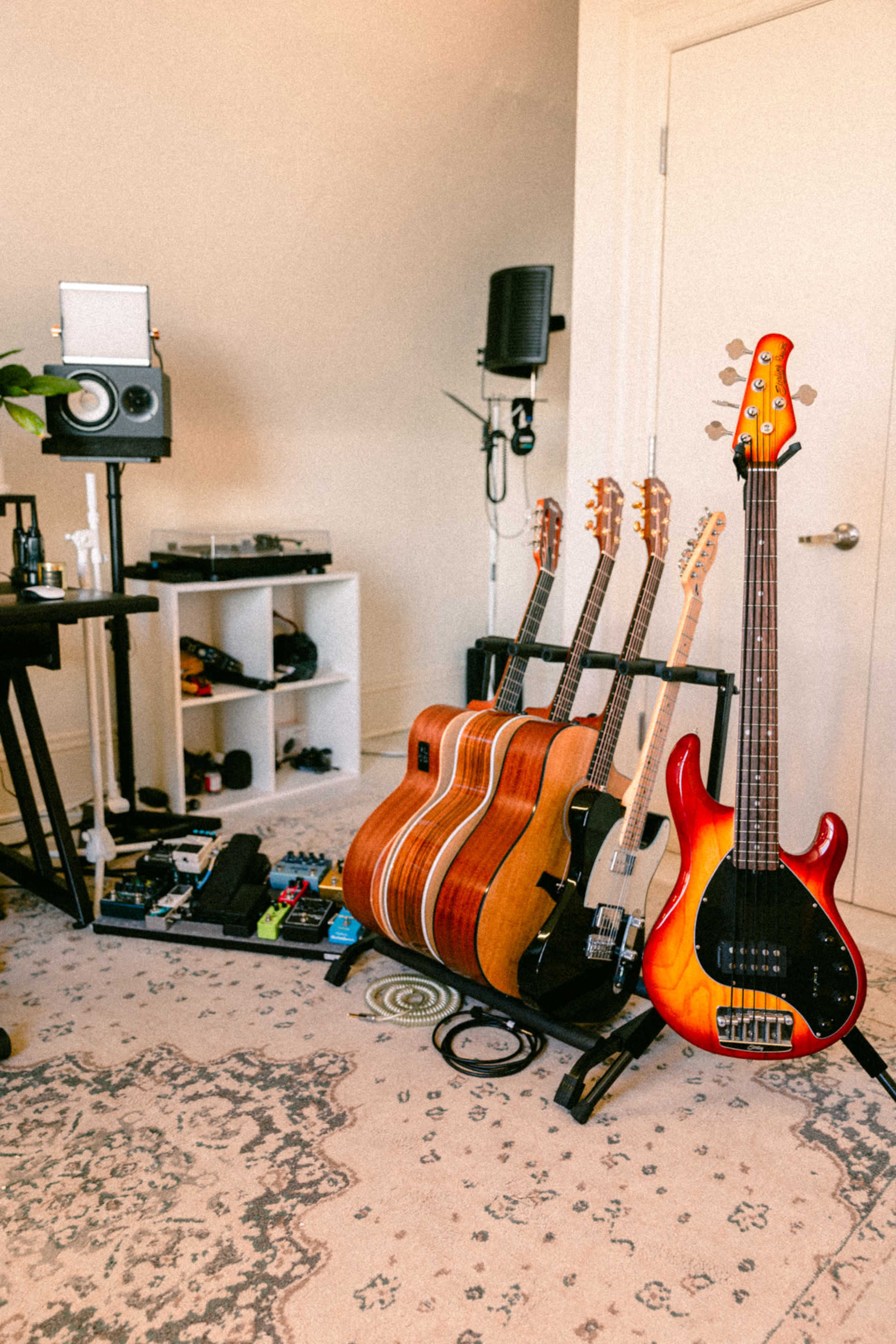 A collection of six guitars, including an electric bass, arranged next to a pedalboard and audio equipment in a music studio.
