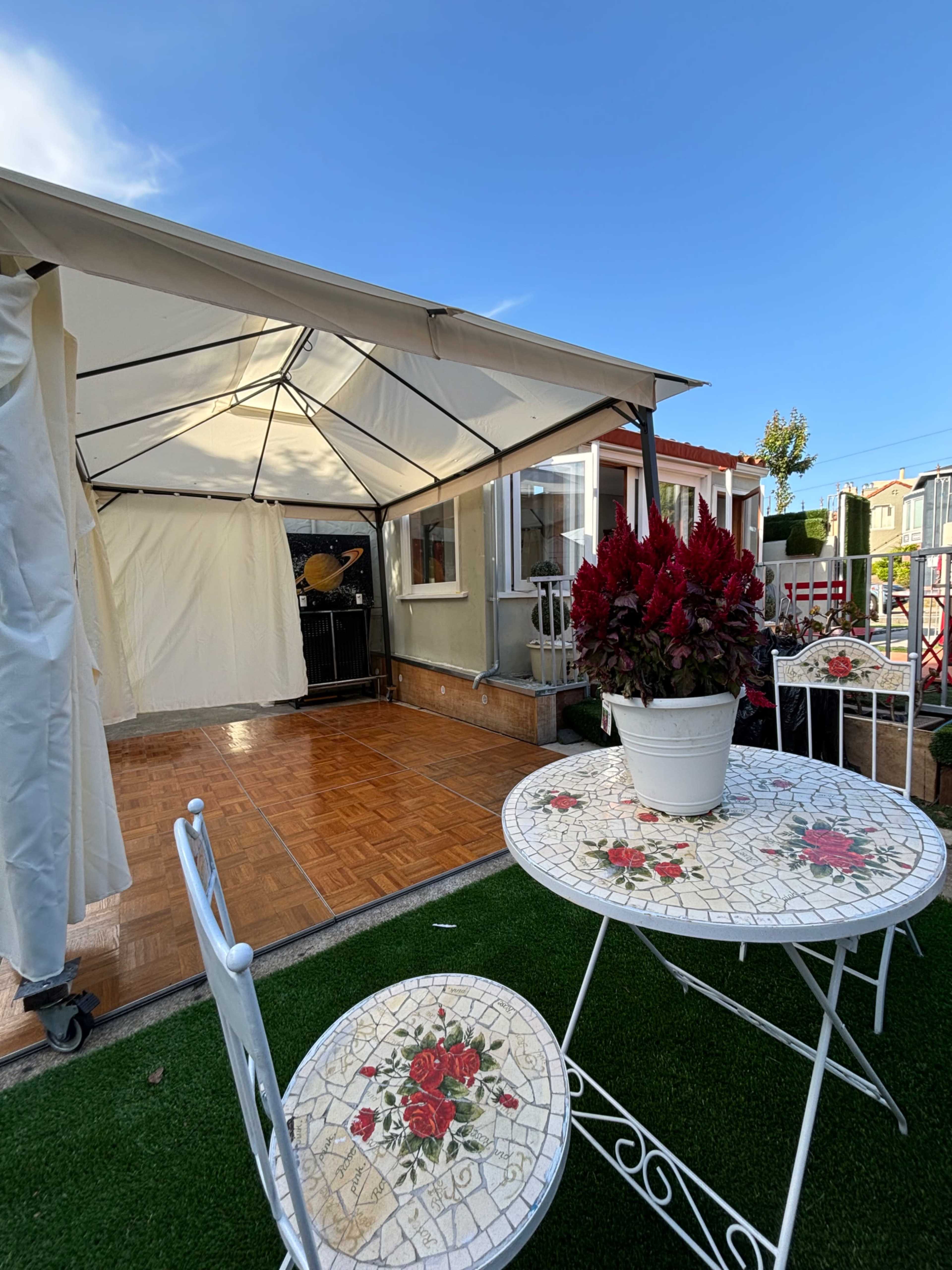 A white canopy covers a small patio area, featuring a round table with floral patterns and two chairs, surrounded by green grass and a potted plant.