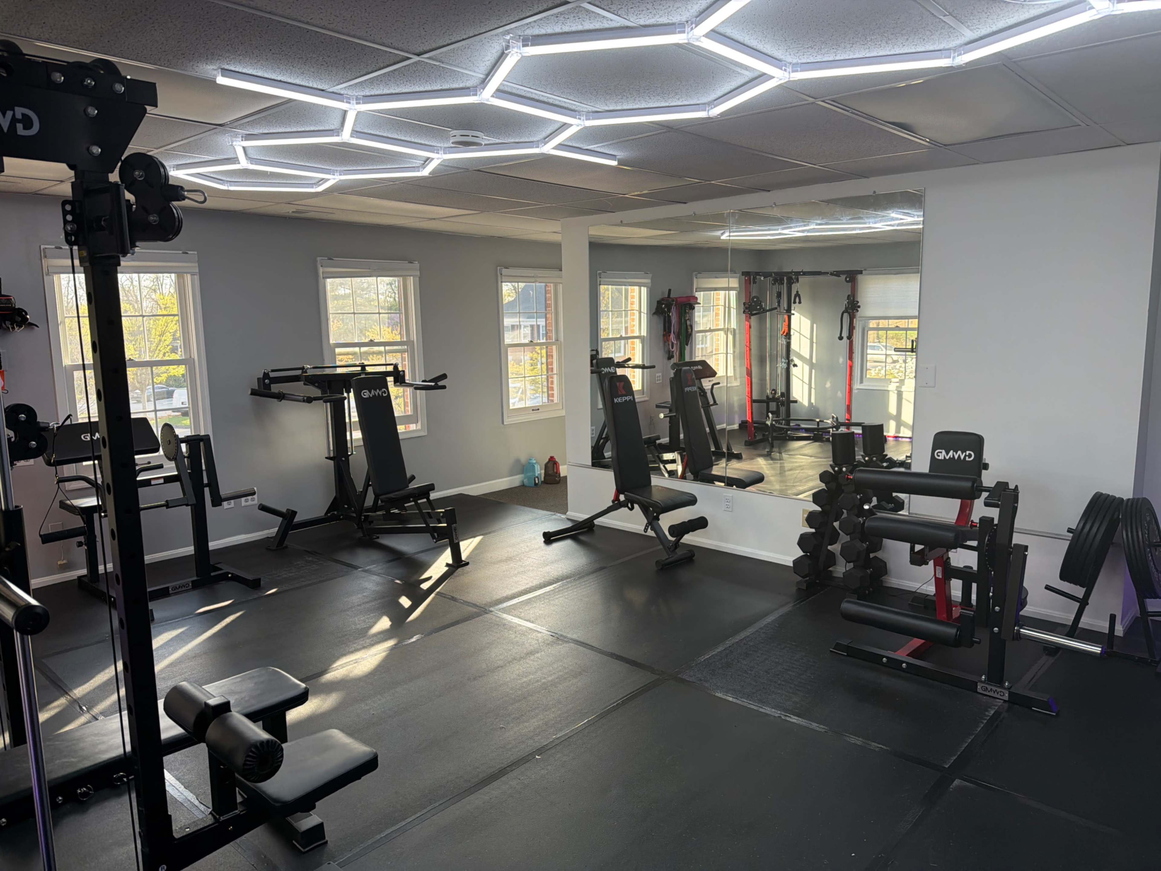 The image shows a modern gym space equipped with various exercise machines and free weights, featuring large windows that allow natural light to enter.