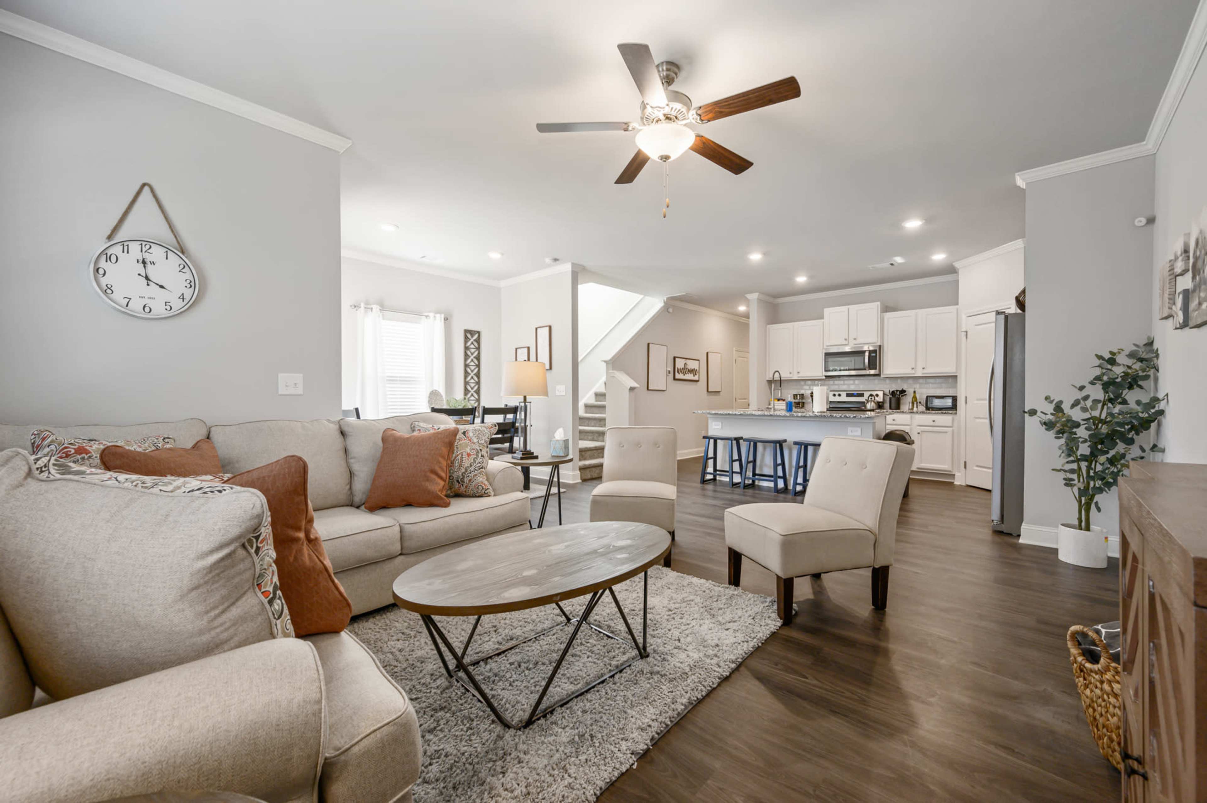 A modern living room with a light-colored sofa, a round coffee table, and a ceiling fan, leading into a kitchen with white cabinets and a dining area.