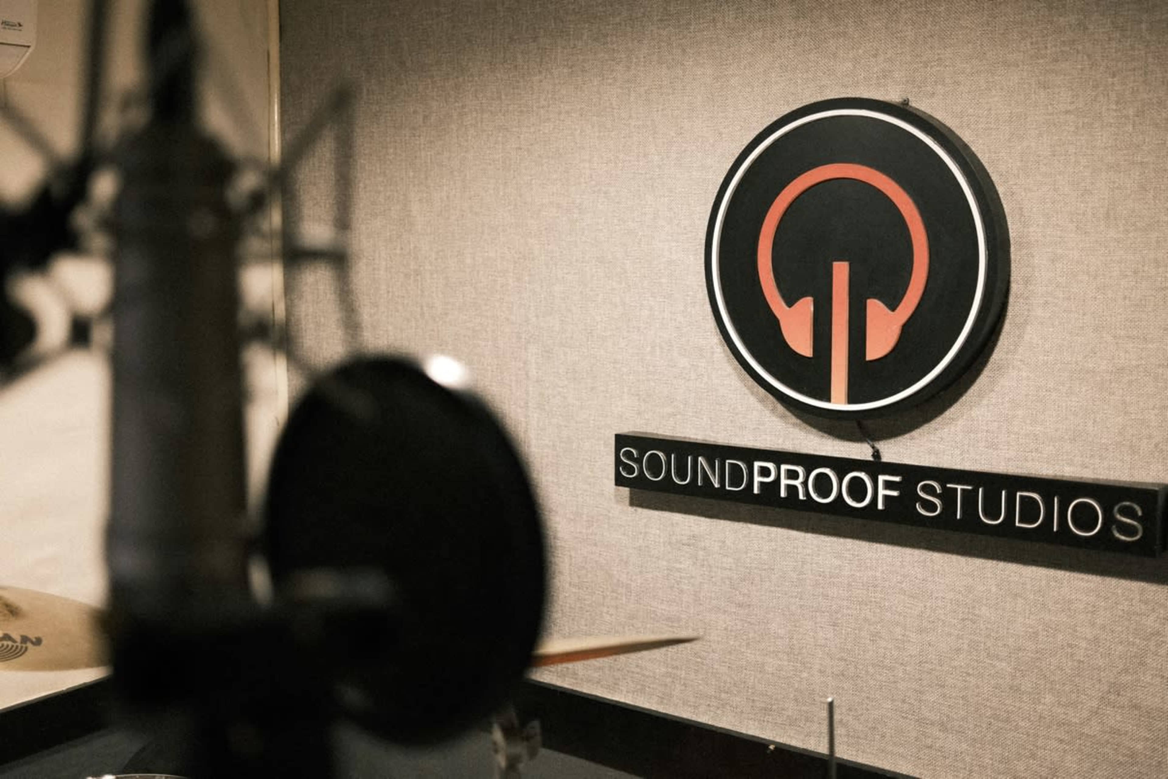 The image shows a recording studio with a microphone in the foreground and a sign for "Soundproof Studios" on a wall in the background.