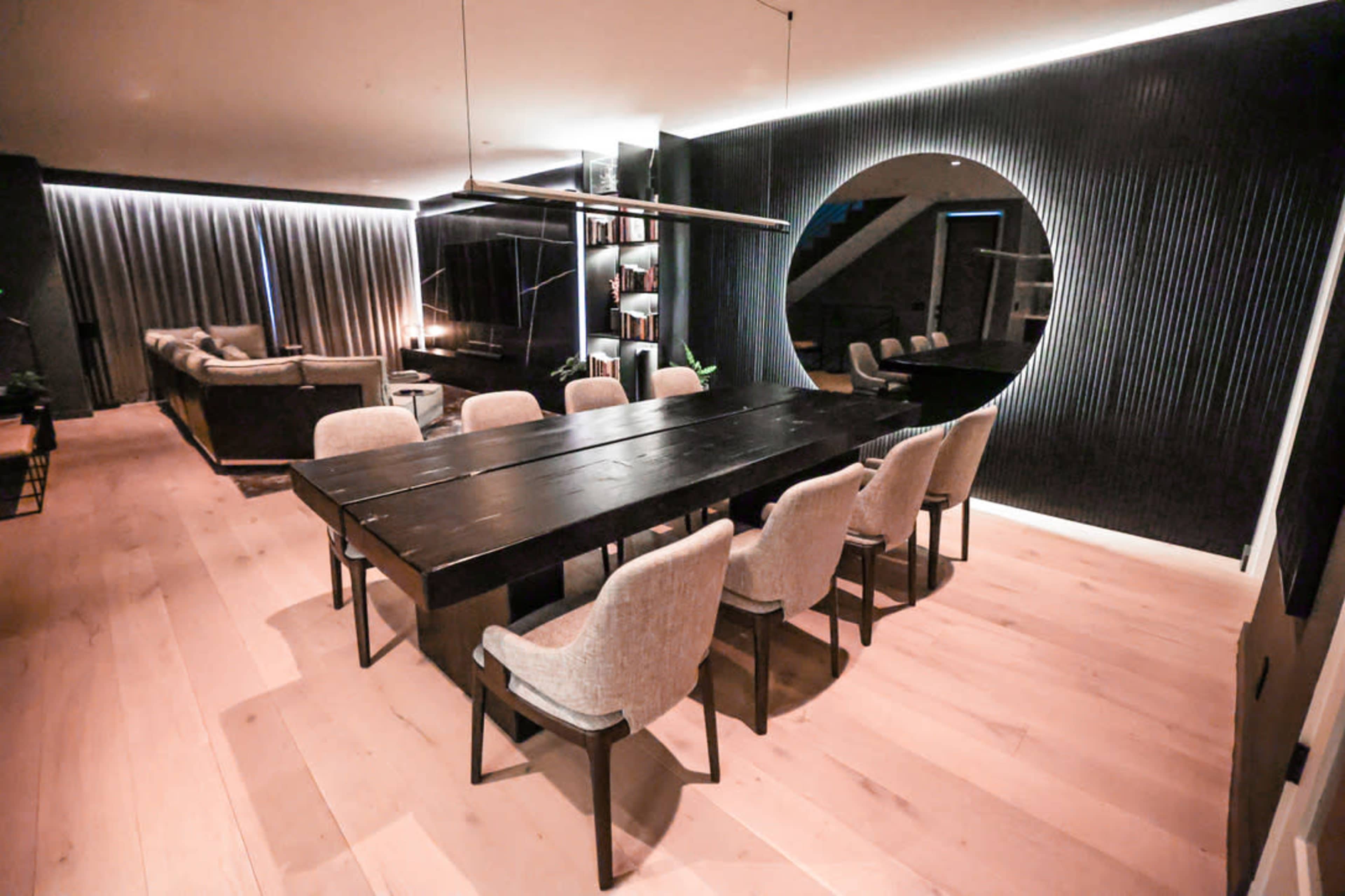 A modern dining room with a long black table surrounded by beige chairs, soft lighting, and a circular mirror on the wall.