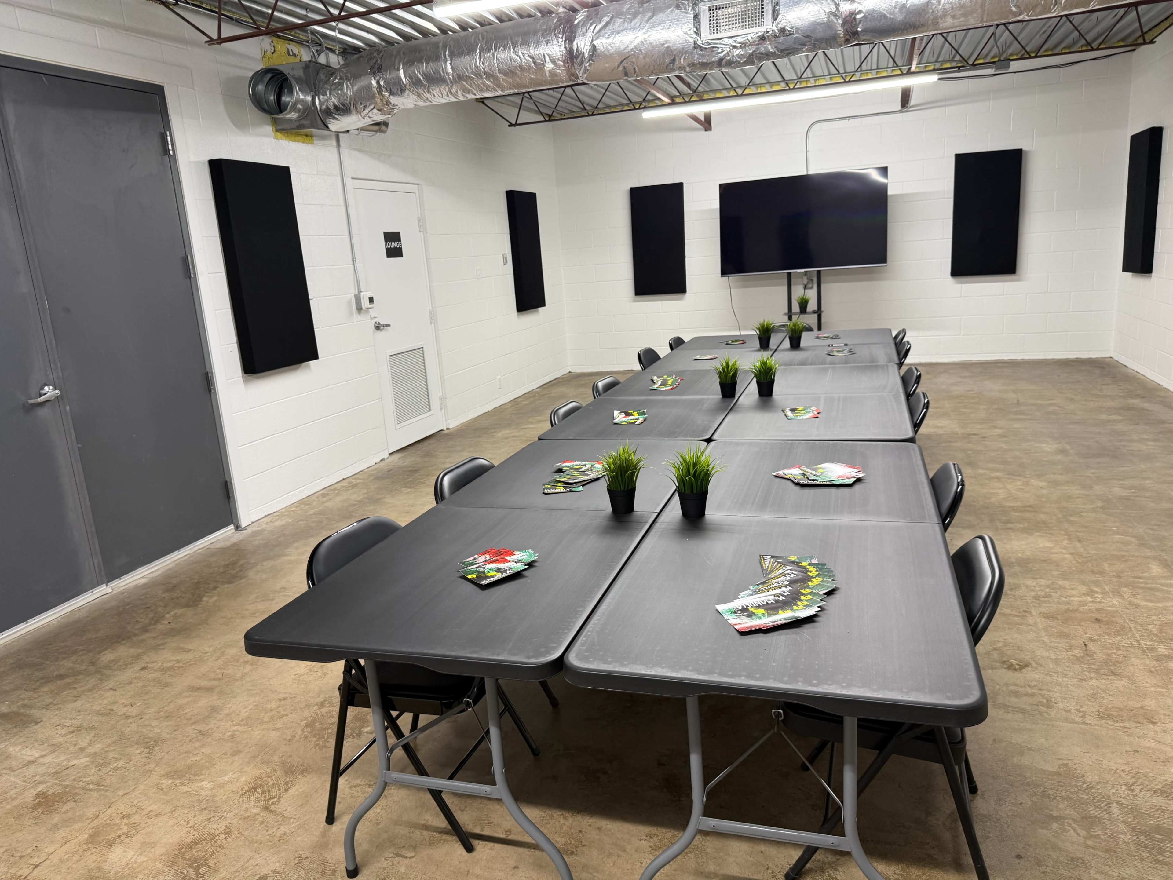 A long rectangular table is set up in a room with white walls, black acoustic panels, and a large screen mounted on one wall.
