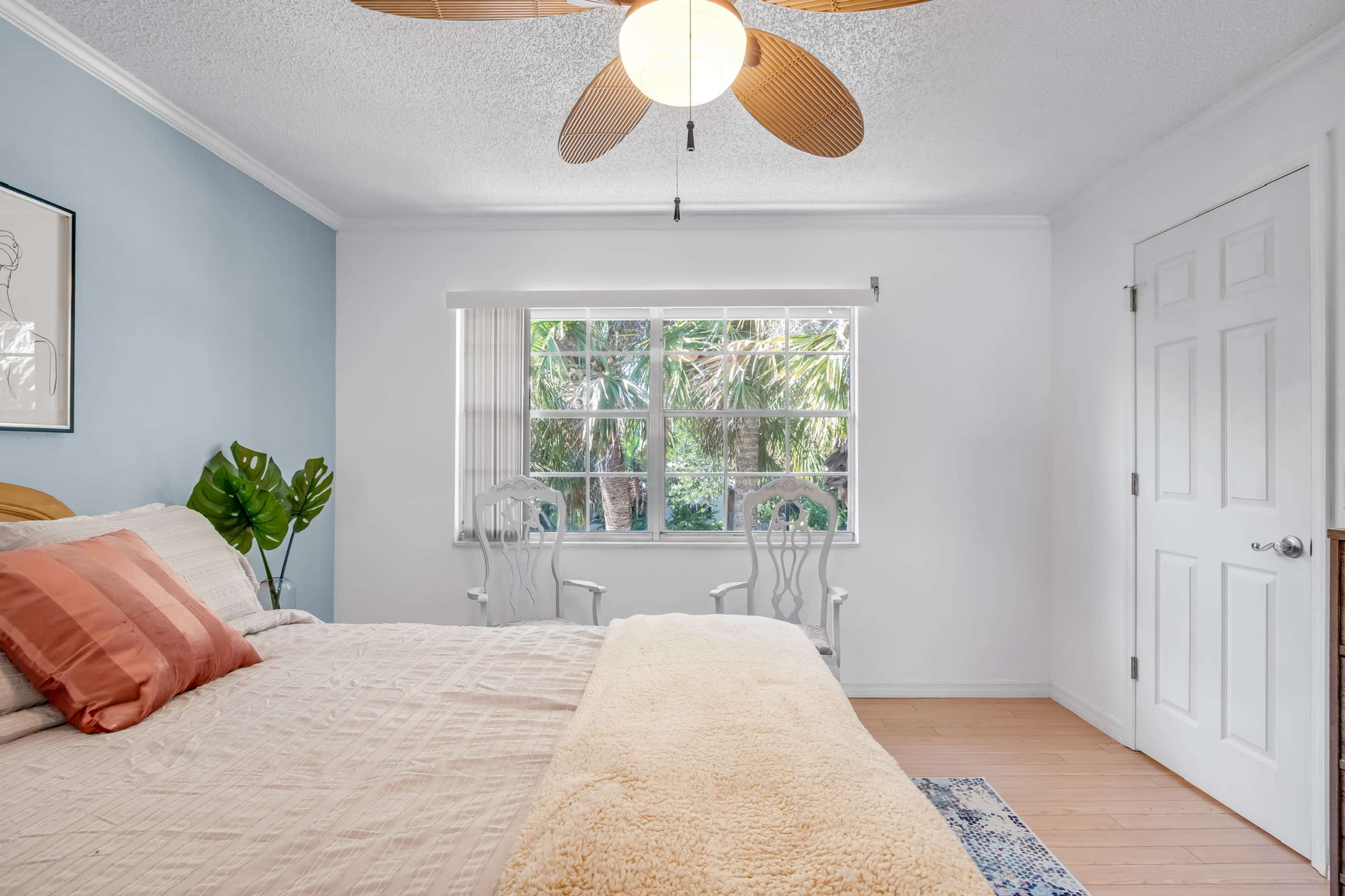A light-colored bedroom features a bed with pillows, two white chairs, and a window overlooking greenery.