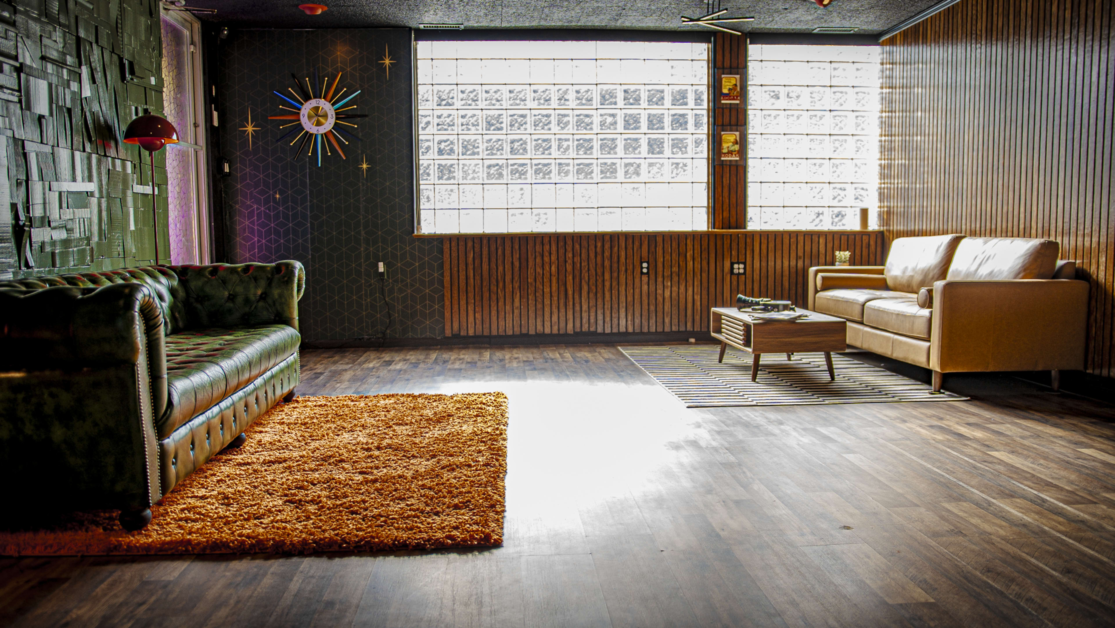 The image shows a spacious room with a green velvet sofa, a brown leather couch, and a wooden coffee table, featuring a large window with glass blocks that allows natural light to enter.